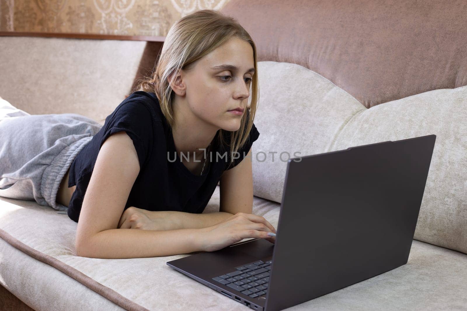 Young woman uses laptop while lying on sofa on her stomach, computer technology for working and studying from home, distance learning and performing work duties