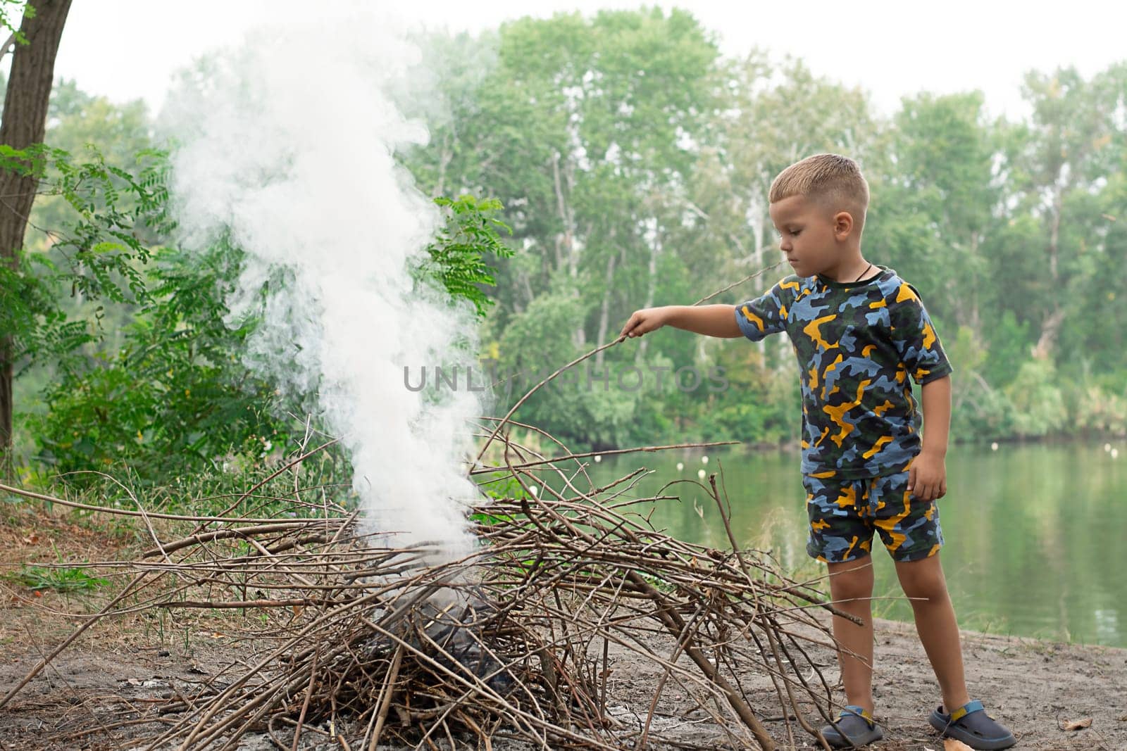 Wildlife concept. Picnic. A little boy, 4 years old, collects dry branches for a fire near a river in the forest in summer. Fishing. Childhood.