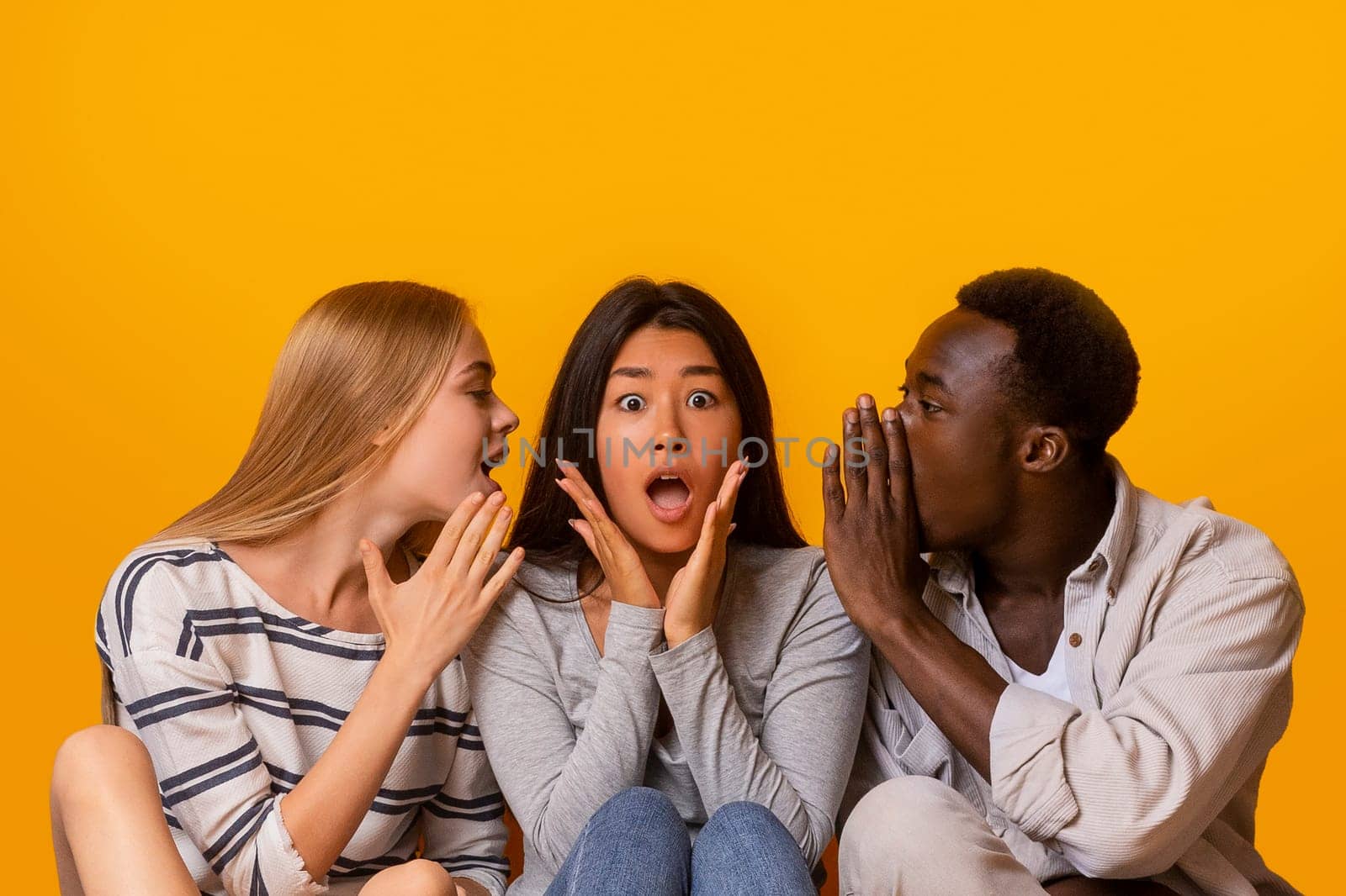 International friends sitting in row on floor, gossiping over yellow studio background, student lifestyle