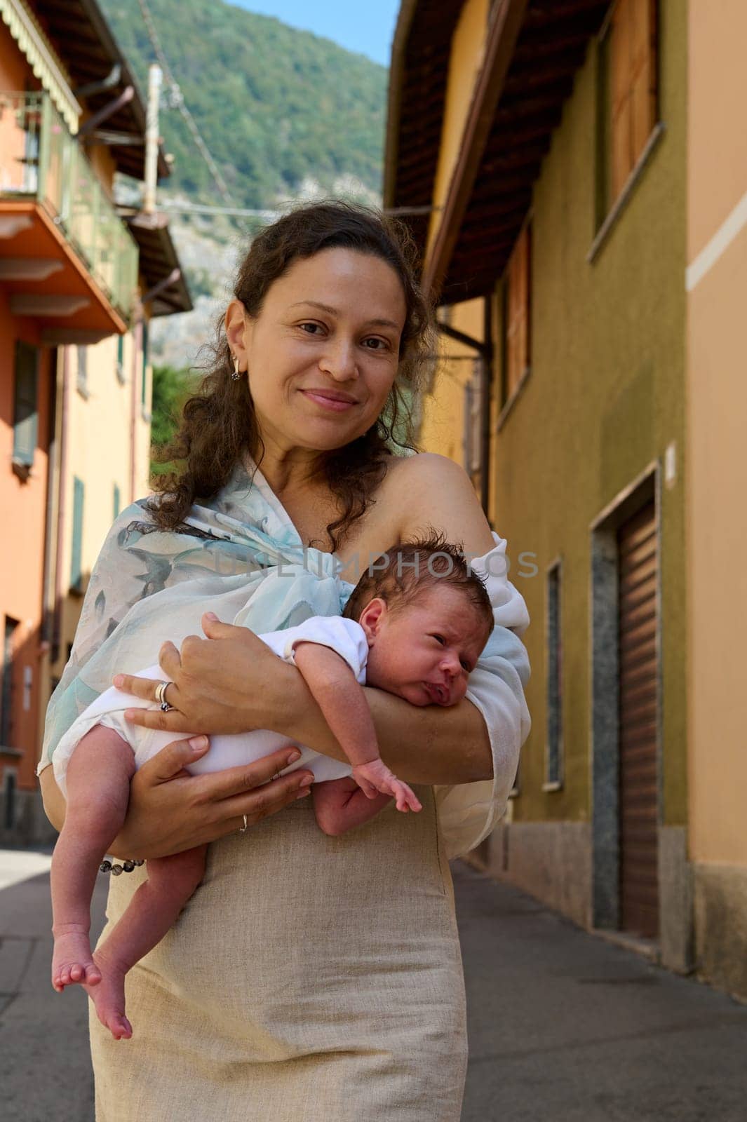 Beautiful woman holding her baby, smiling looking at camera, standing in cobblestone alley in Italian medieval village by artgf