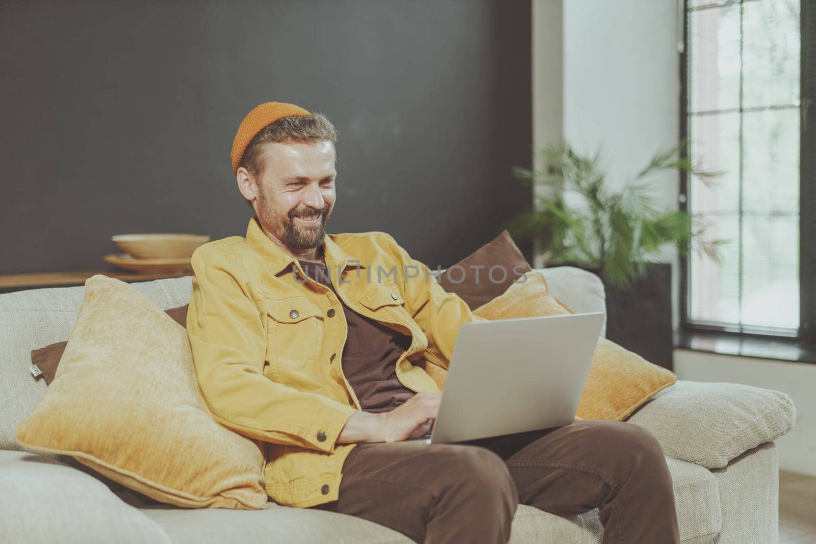 Smiling young man enjoys comfort of home while working on laptop. Laptop on knees, exemplifies modern lifestyle of remote work and telecommuting, showcasing perfect balance between work and relaxation by LipikStockMedia