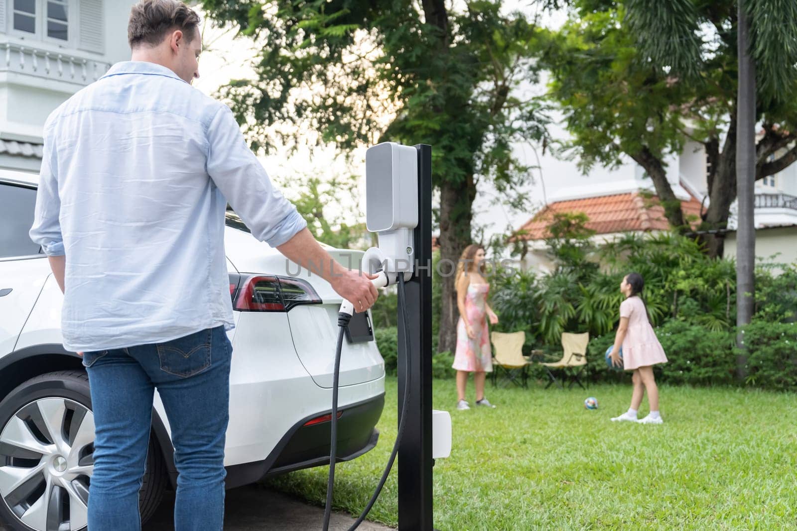 Modern and environmental conscious man recharge electric vehicle from home charging station on while his playful family playing together in background. Energy sustainable for better future. Synchronos