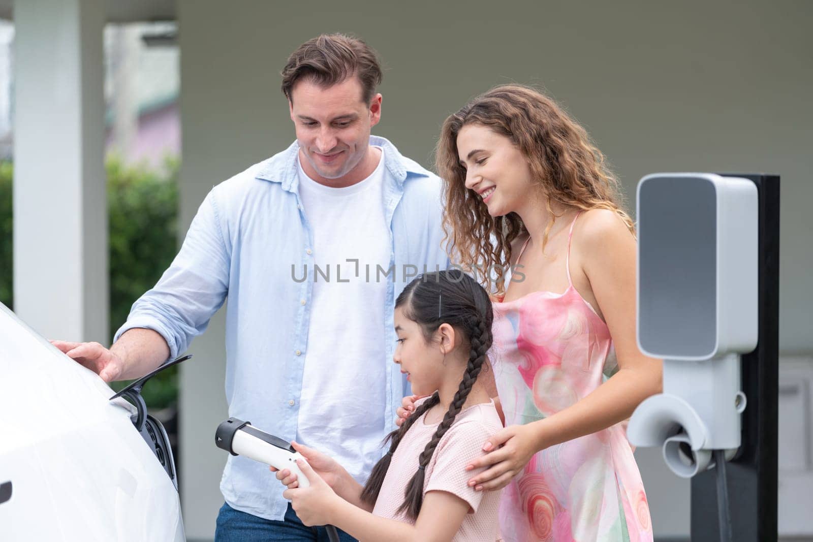 Happy little young girl learn about eco-friendly and energy sustainability as she help her family recharge electric vehicle from home EV charging station. EV car and modern family concept. Synchronos