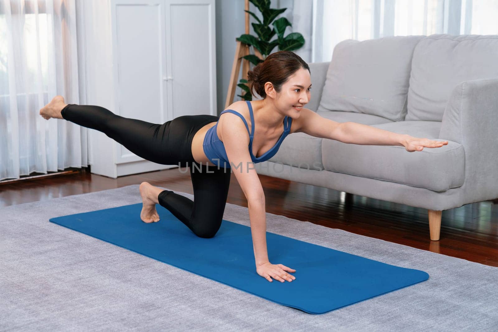 Asian woman in sportswear doing yoga exercise on fitness mat as her home workout training routine. Healthy body care and calm meditation in yoga lifestyle with comfortable and relaxation. Vigorous