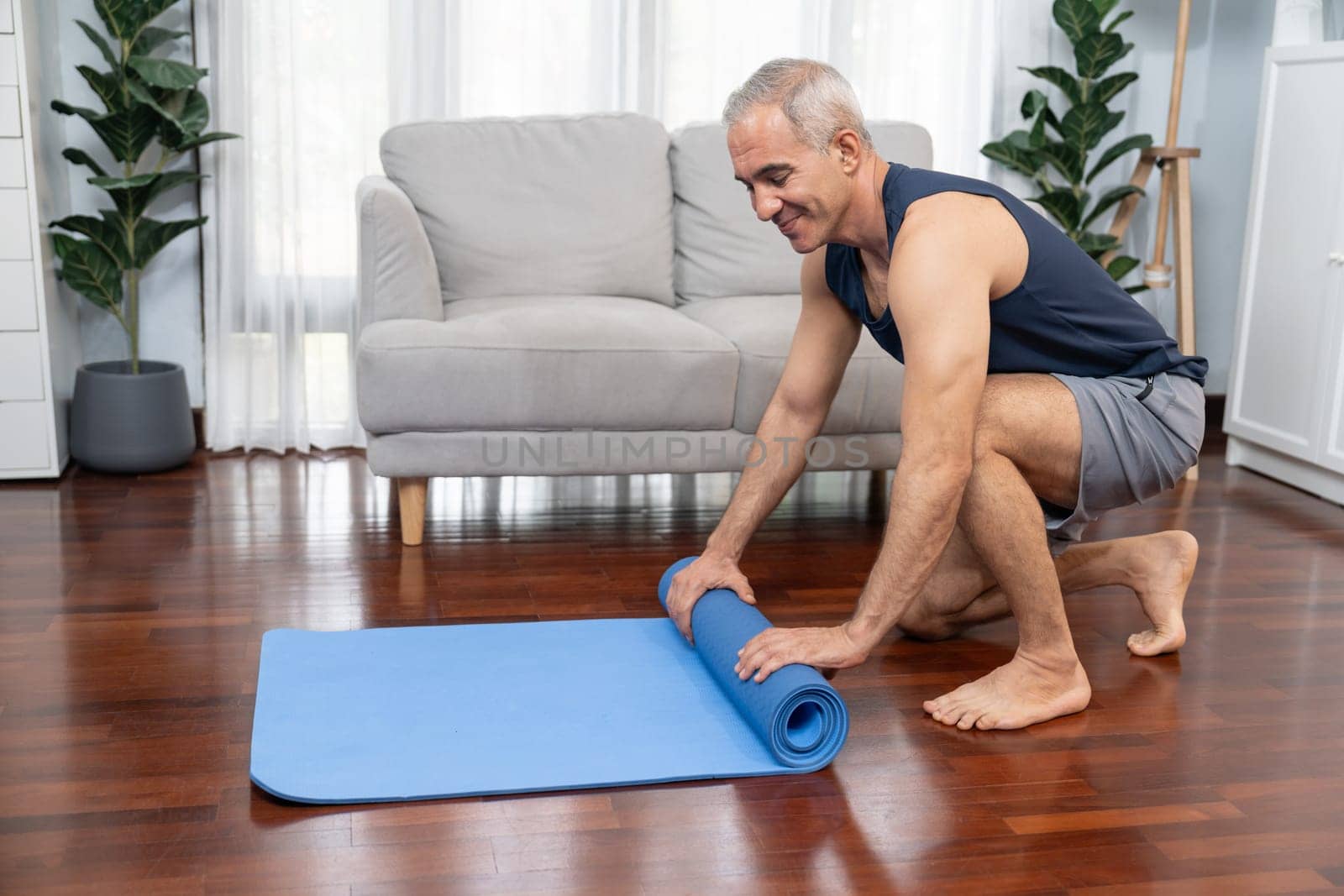 Active and sporty senior man preparing, rolling fitness exercising mat on living room floor at home. Home exercise as concept of healthy fit body lifestyle after retirement. Clout