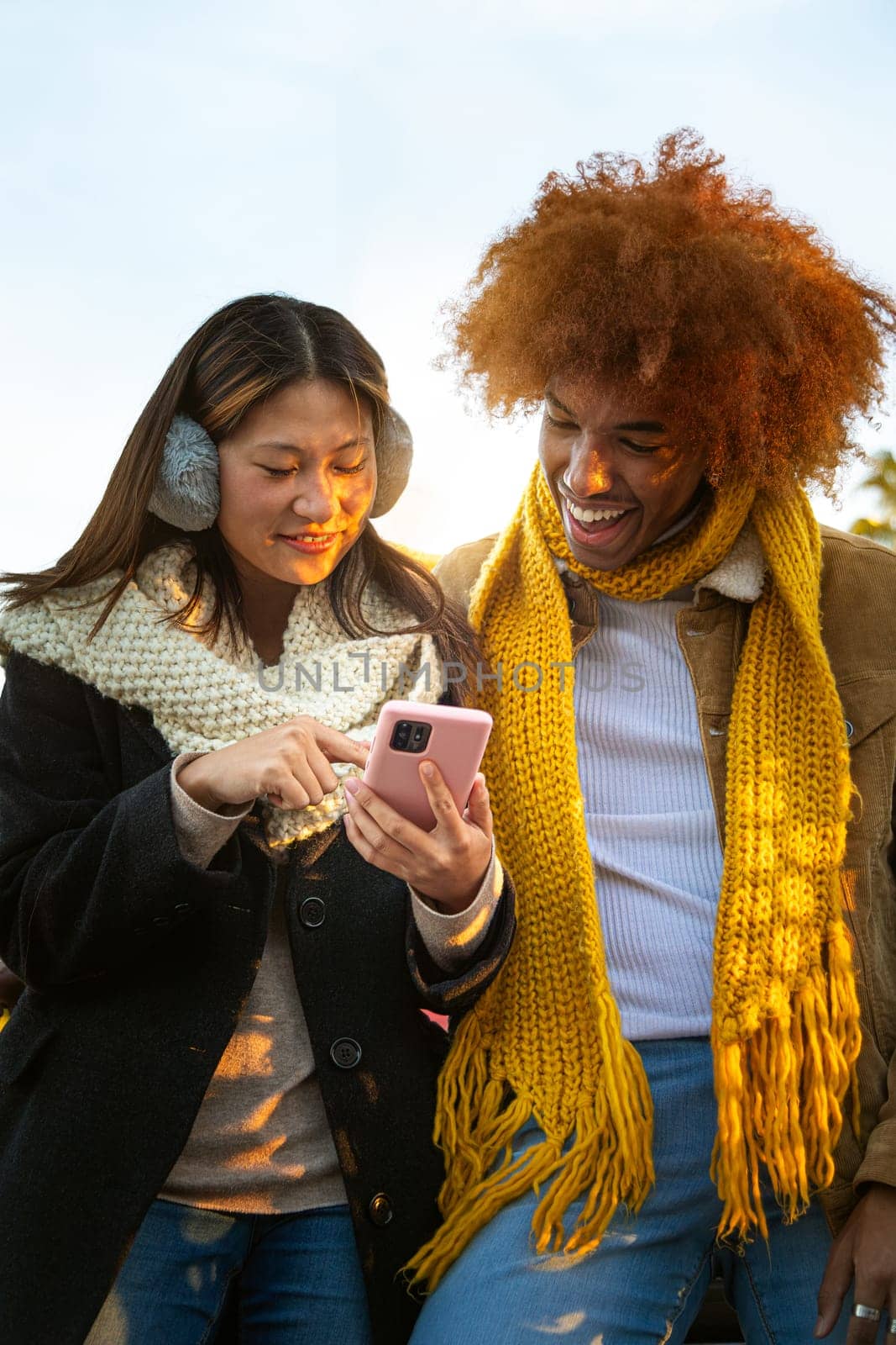 Vertical portrait of happy multiracial young couple looking smartphone together outdoors on a winter day. Multiethnic friends looking social media apps together. Technology concept.