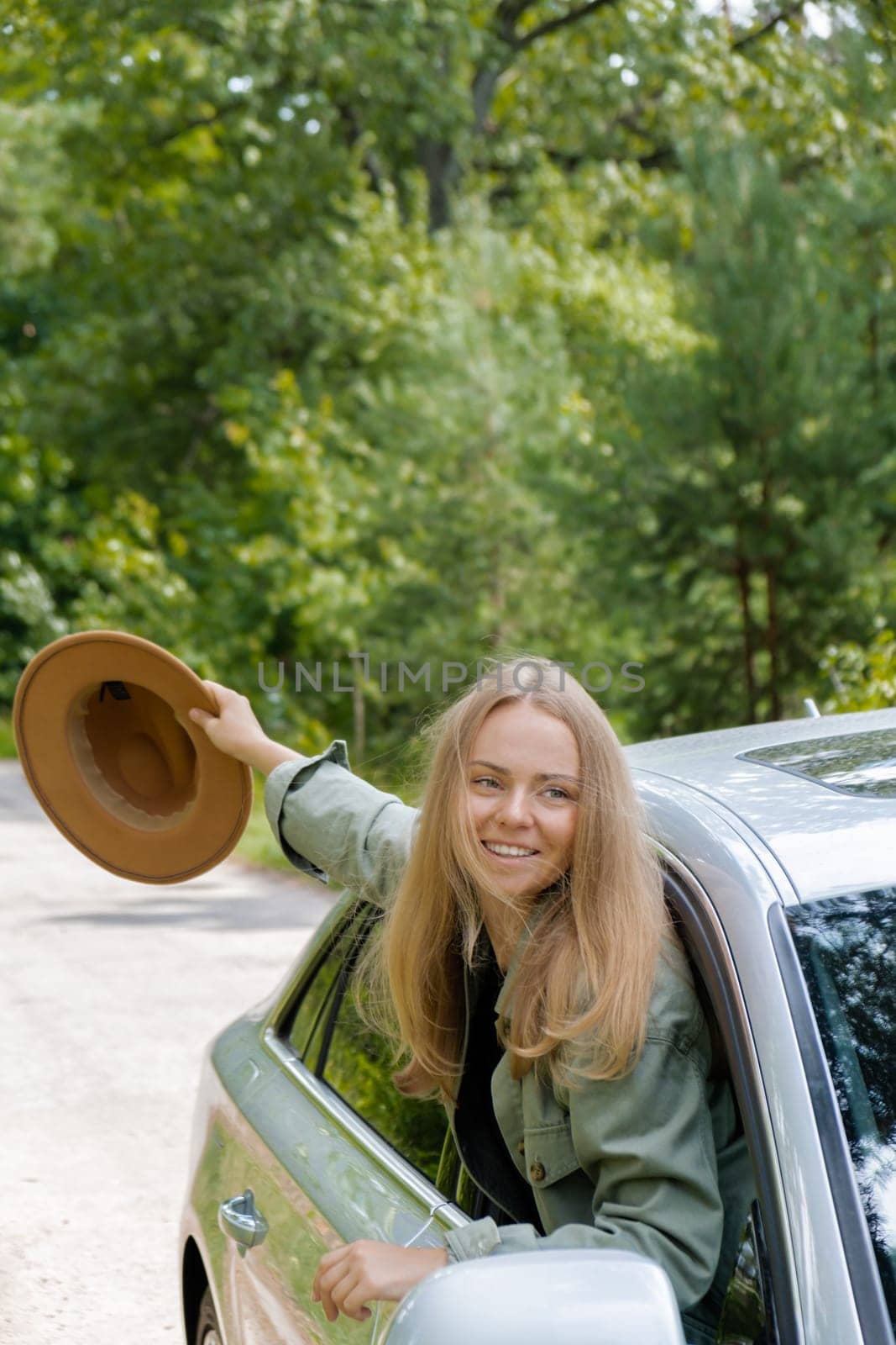 Smiling young woman in hat looking from car window. Local solo travel on weekends concept. Exited woman explore freedom outdoors in forest. Unity with nature lifestyle, rest recharge relaxation