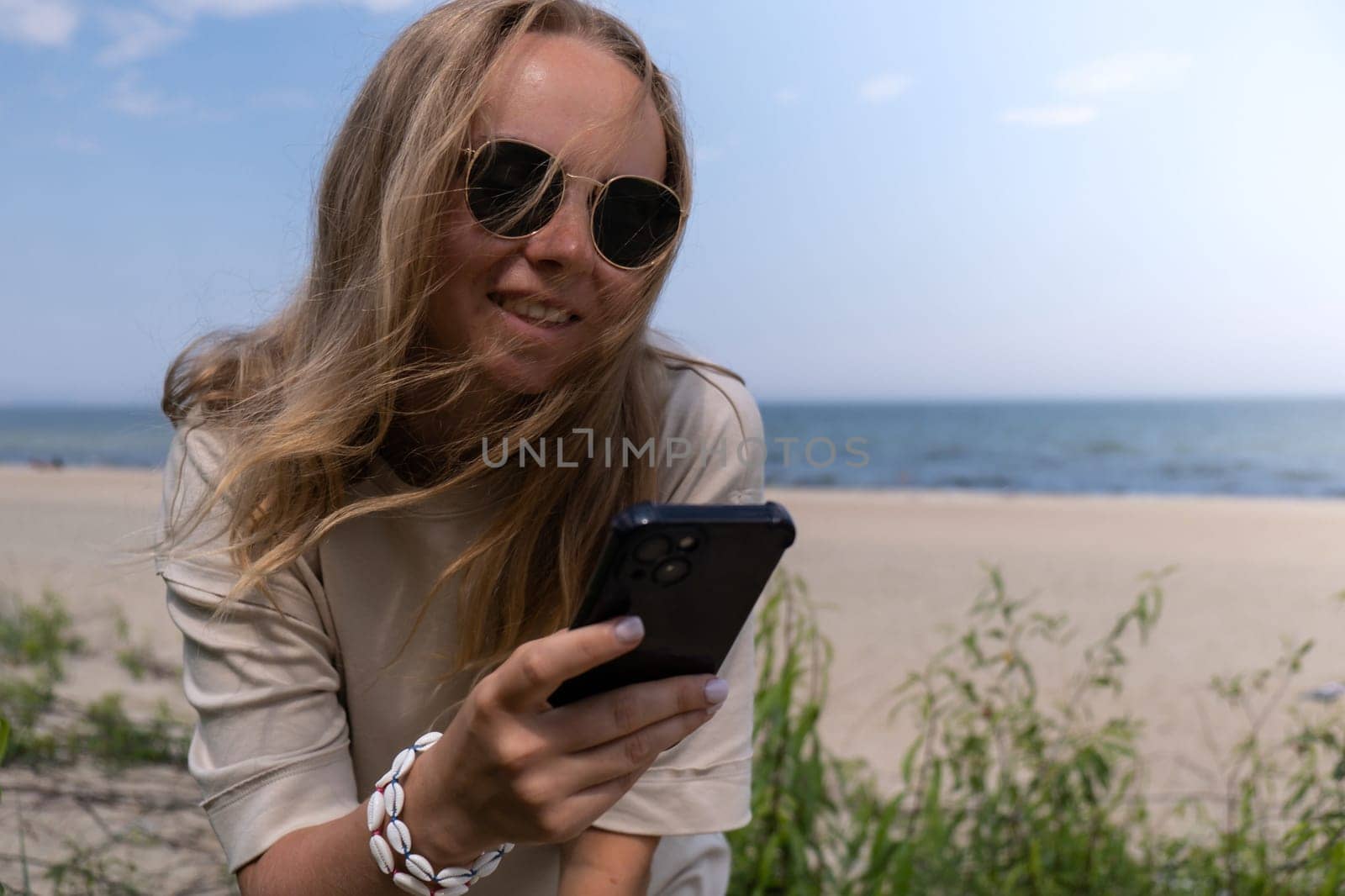Young woman in sunglasses with shell bracelets holding smartphone. Influencer Using mobile phone for video call selfie waving on the beach seacoast. Holiday vacation advertisement concept. Social media