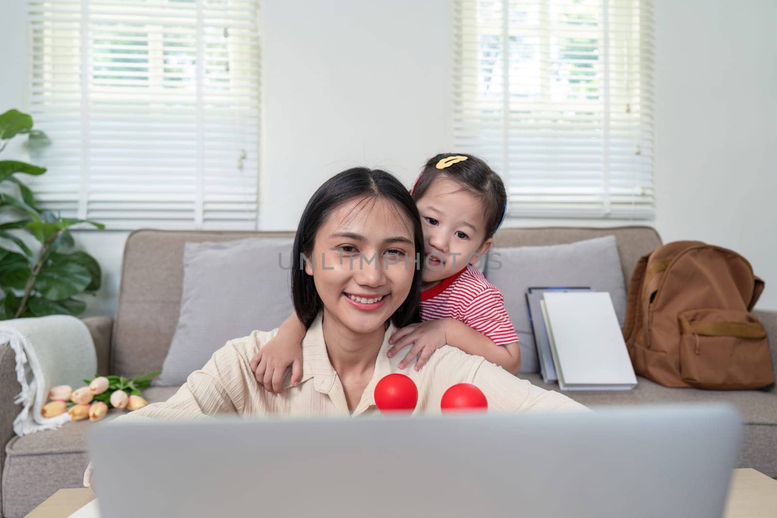 A mother and her young daughter smiling and using a laptop in a bright, cozy living room with natural light streaming through the windows.