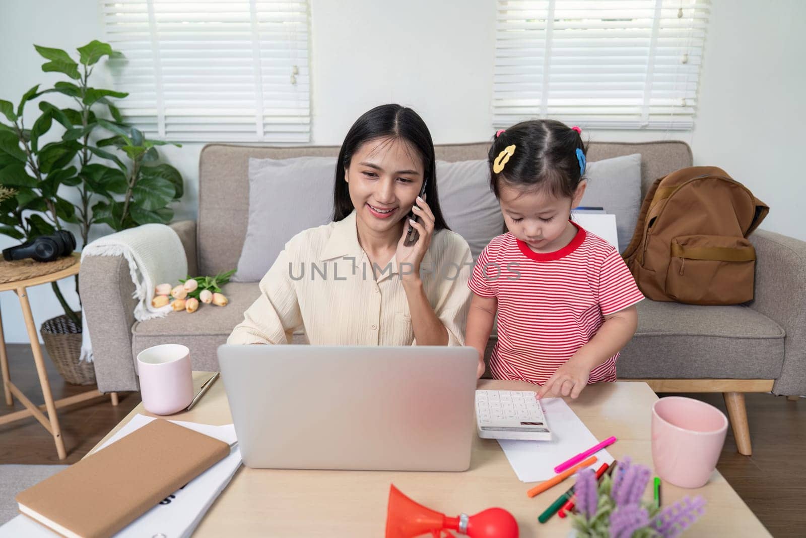 A mother multitasking with a laptop and phone while her daughter plays beside her in a cozy living room setting.
