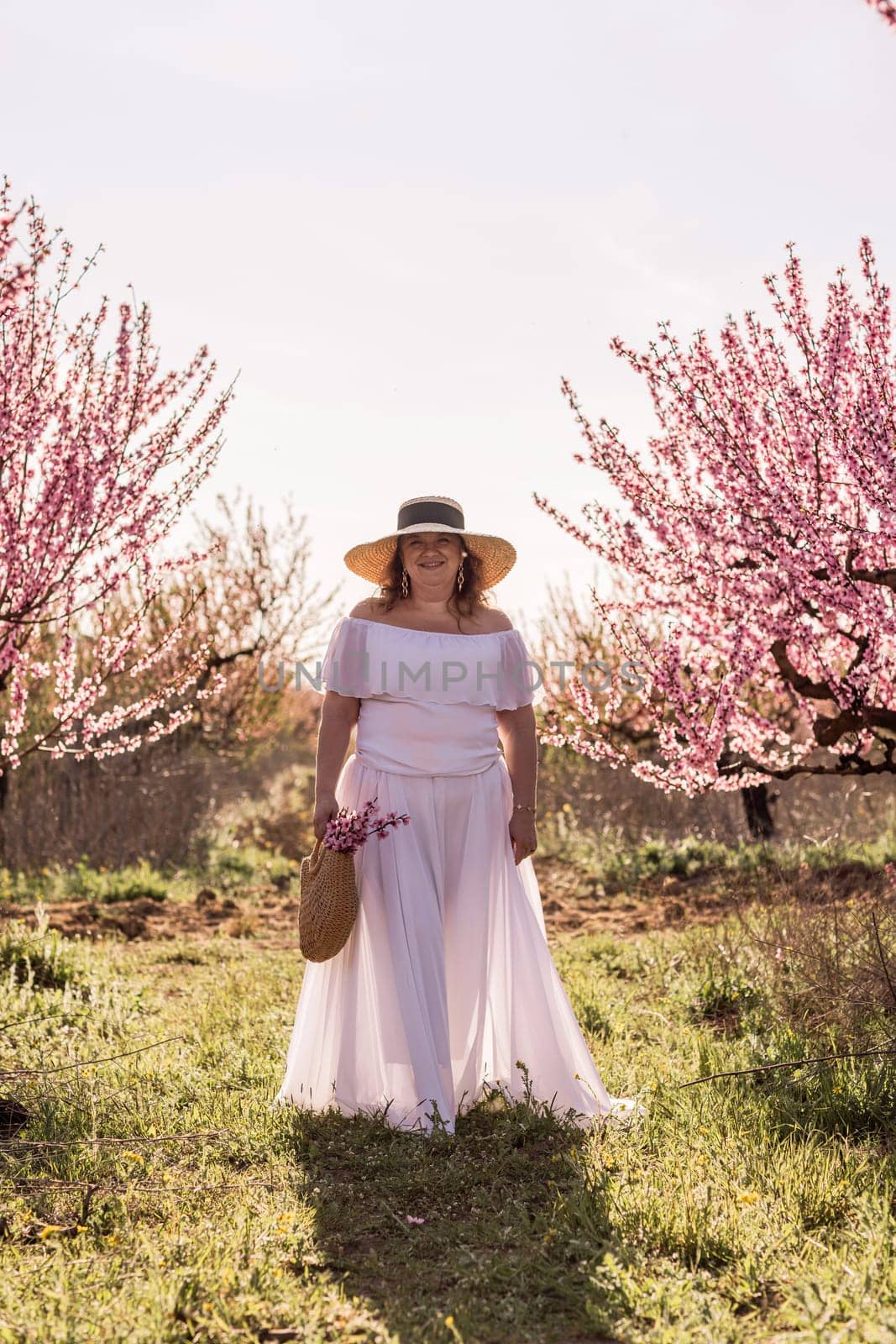 Woman blooming peach orchard. Against the backdrop of a picturesque peach orchard, a woman in a long white dress and hat enjoys a peaceful walk in the park, surrounded by the beauty of nature