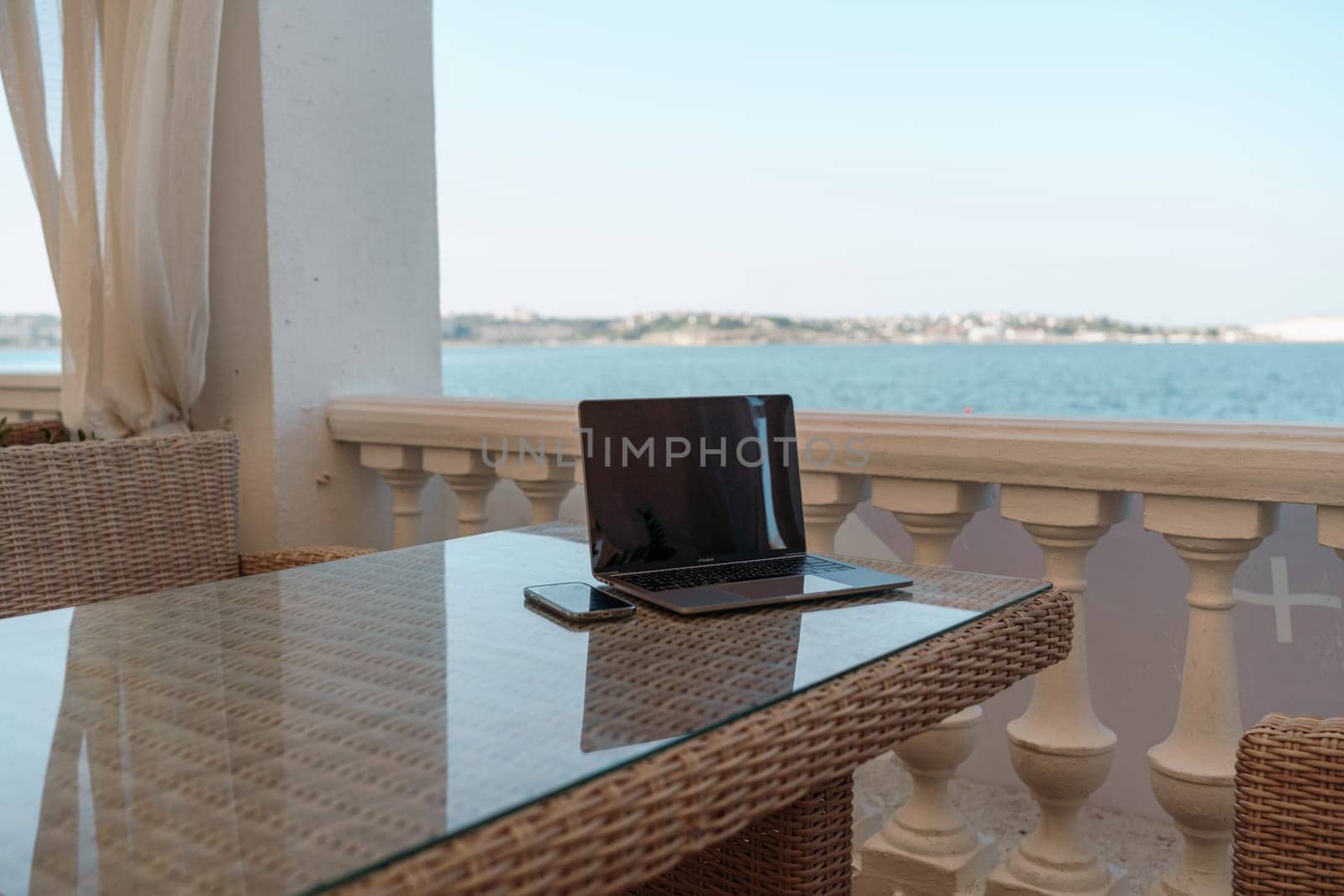 A laptop and a cell phone are on a table by a beach. The scene is peaceful and relaxing, with the ocean in the background. The table is made of clear plastic