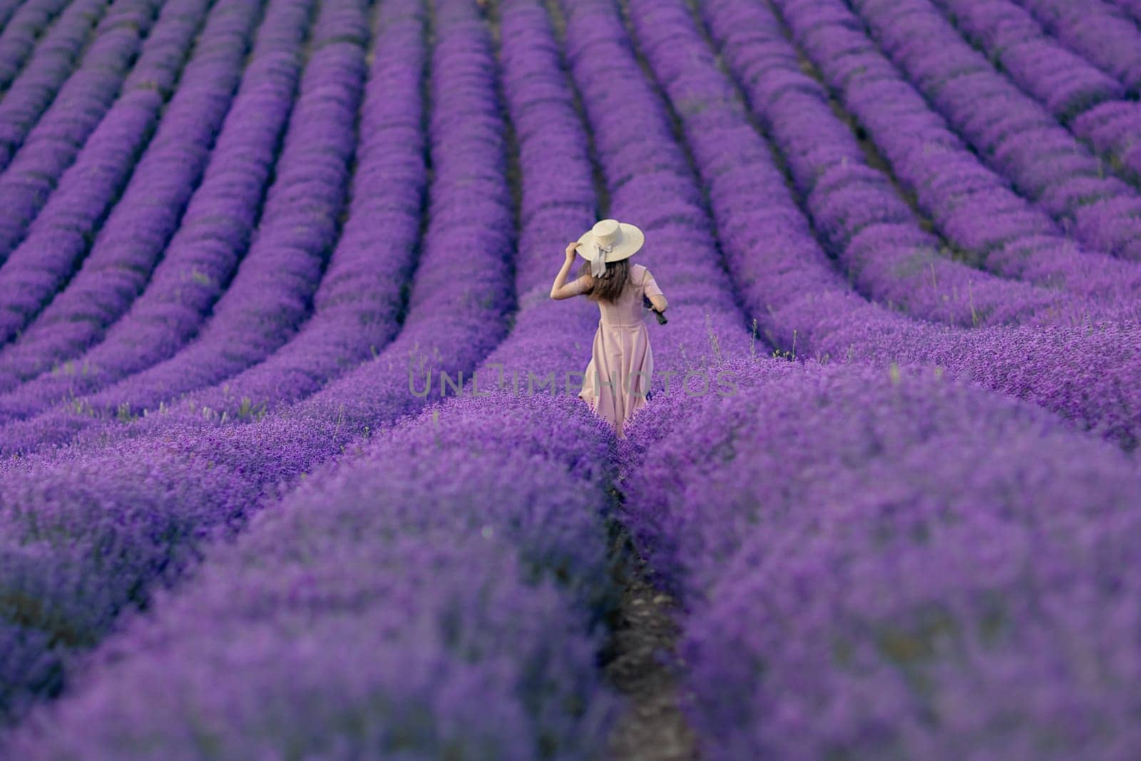 A woman is walking through a field of purple flowers. The field is vast and stretches out in front of her. The woman is wearing a white hat and a pink dress