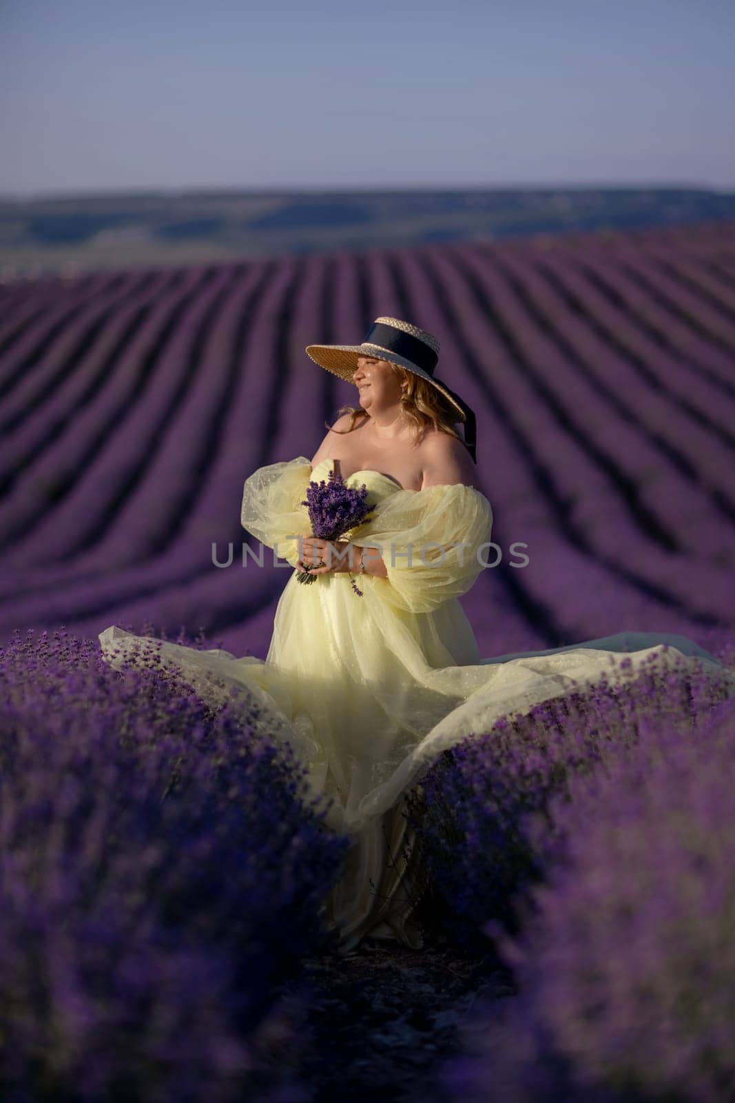 Woman poses in lavender field at sunset. Happy woman in yellow dress holds lavender bouquet. Aromatherapy concept, lavender oil, photo session in lavender.