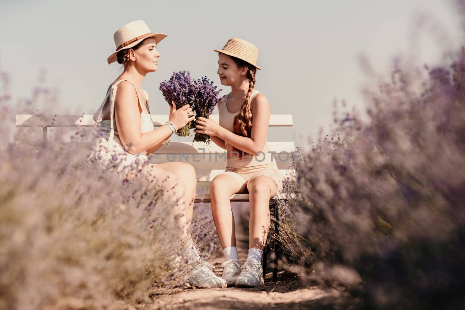 A woman and a child are sitting on a bench in a field of purple flowers lavande. The woman is holding a bouquet of flowers and the child is holding a bouquet of flowers as well.