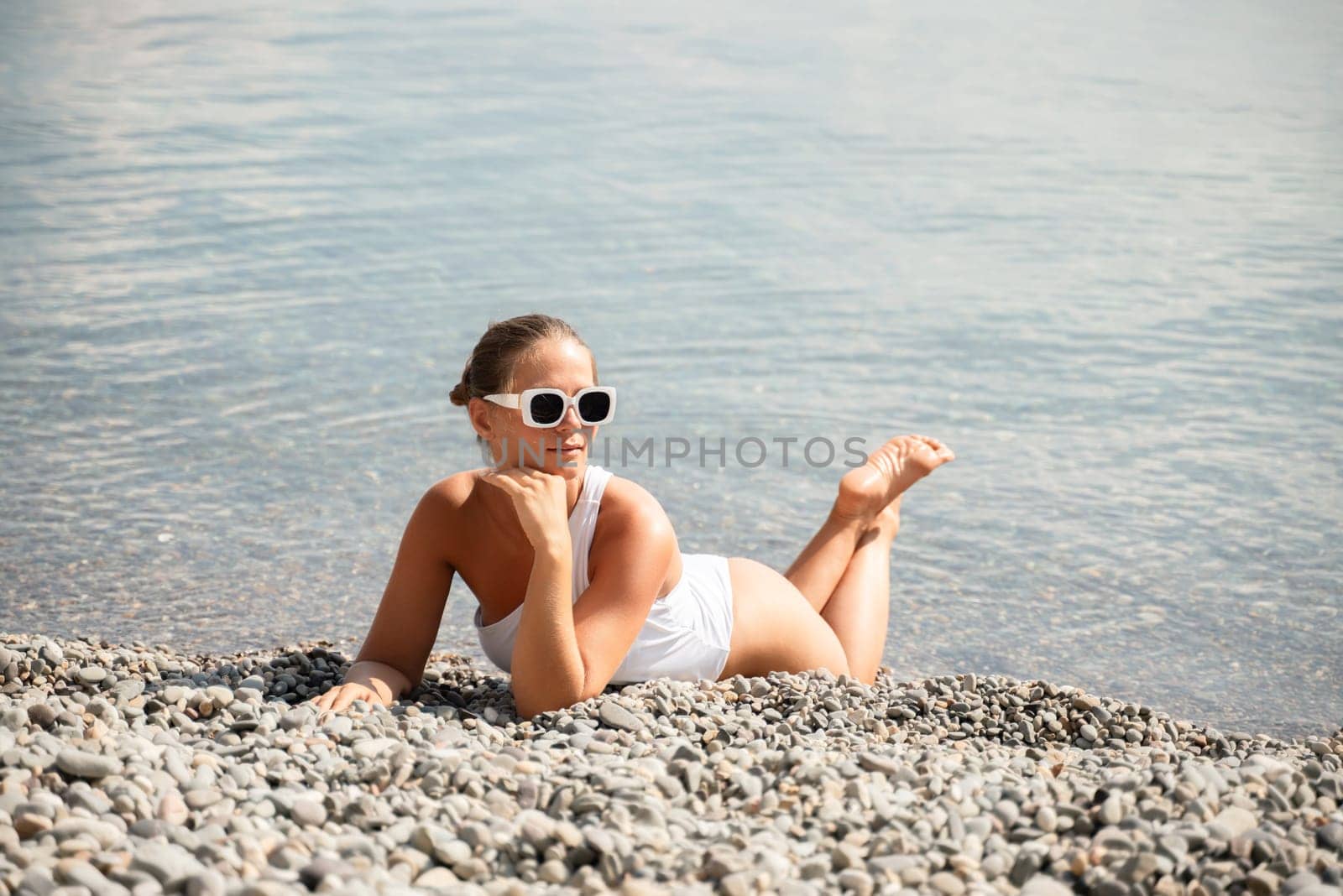 A woman is laying on the beach wearing sunglasses and a white tank top. She is looking at the camera