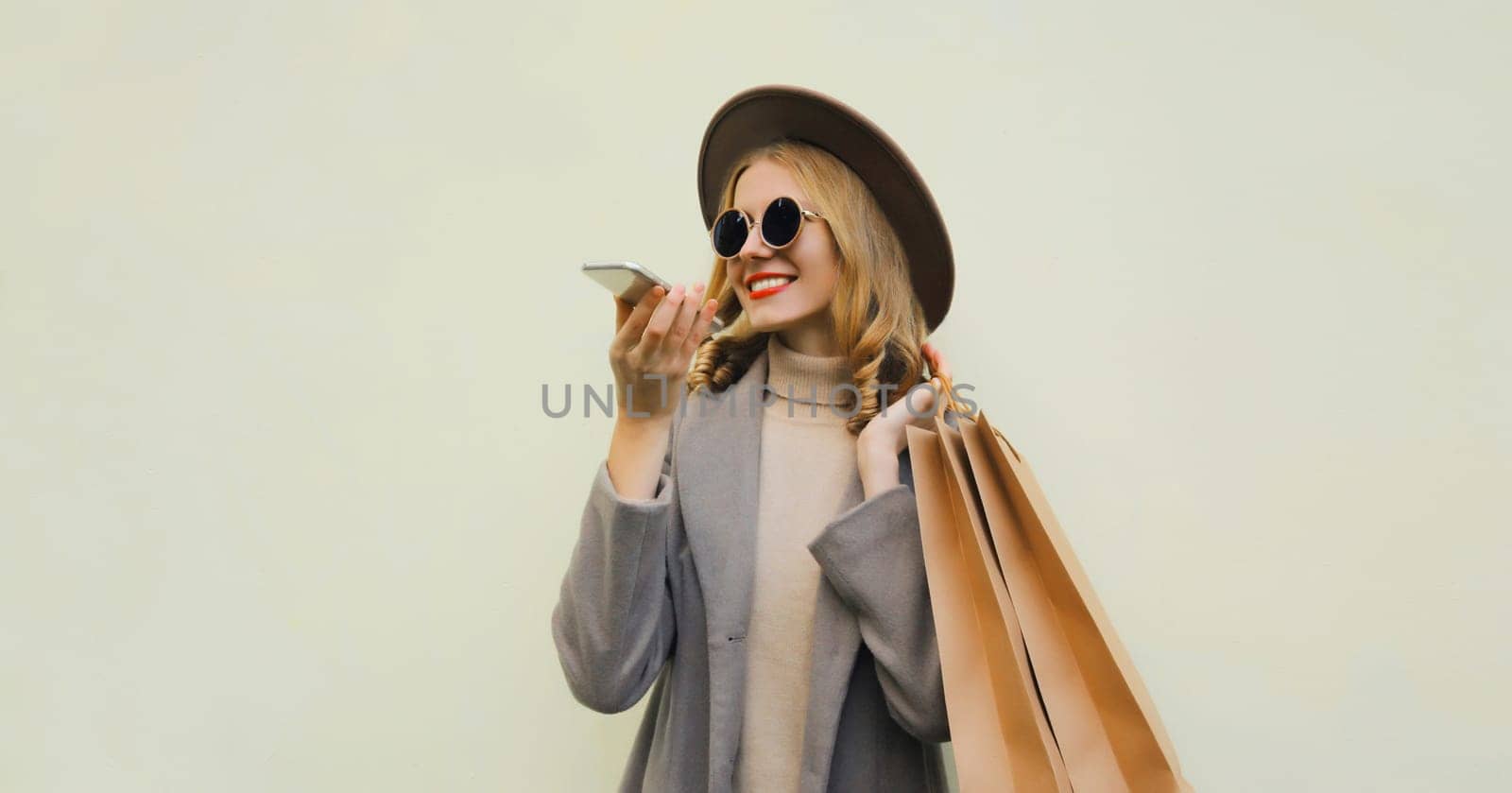 Beautiful happy young woman calling on smartphone with shopping bag, pretty girl smiles wearing hat, glasses standing on brown background