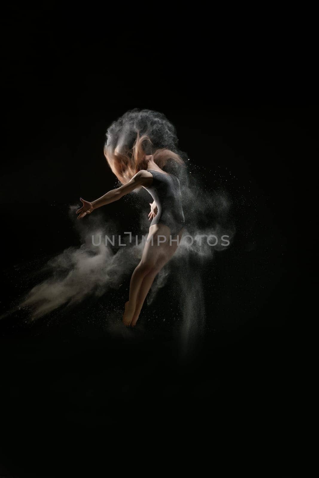 Female sexy gymnast in gray bodysuit shot in a jump on black background in a cloud of white dust all over body