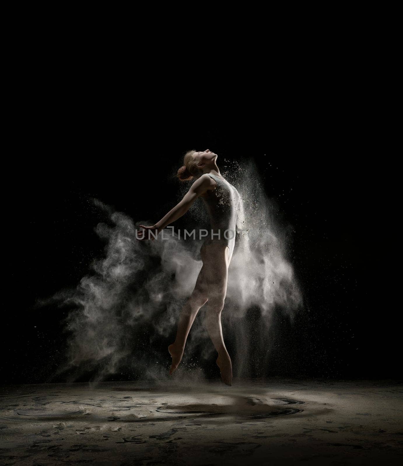 Female sexy gymnast in bodysuit shot in a jump on black background in a cloud of white dust all over body