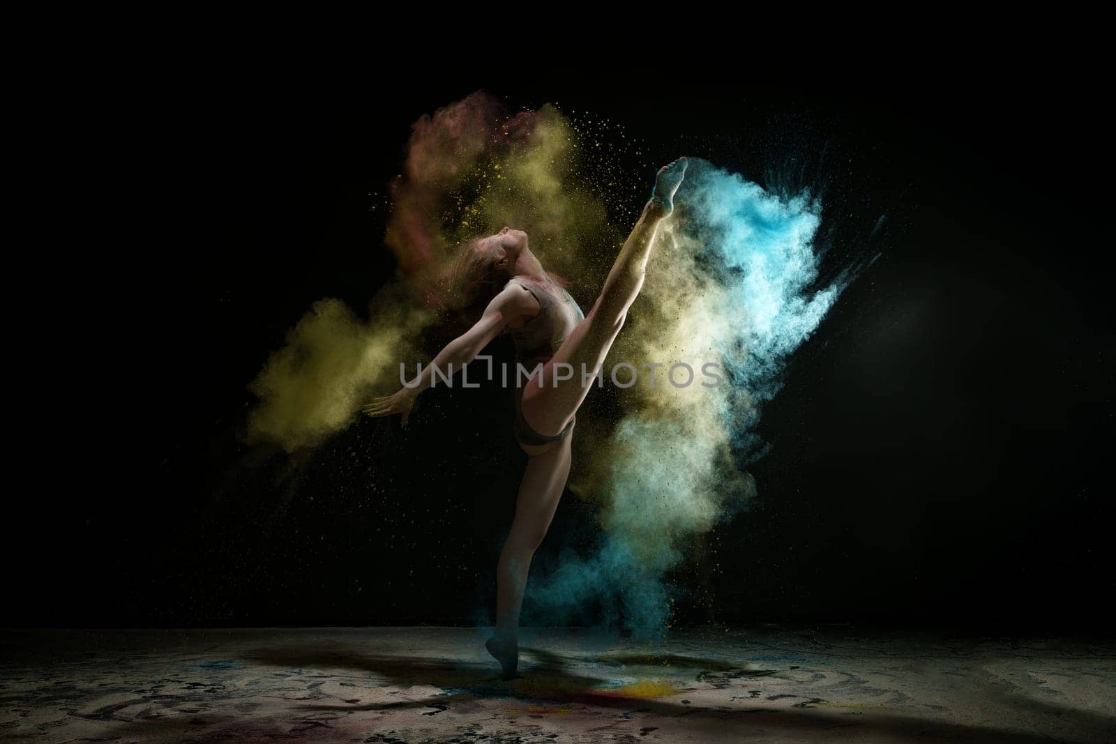 Female sexy gymnast in gray bodysuit shot in a jump on black background in a cloud of blue and yellow dust all over body