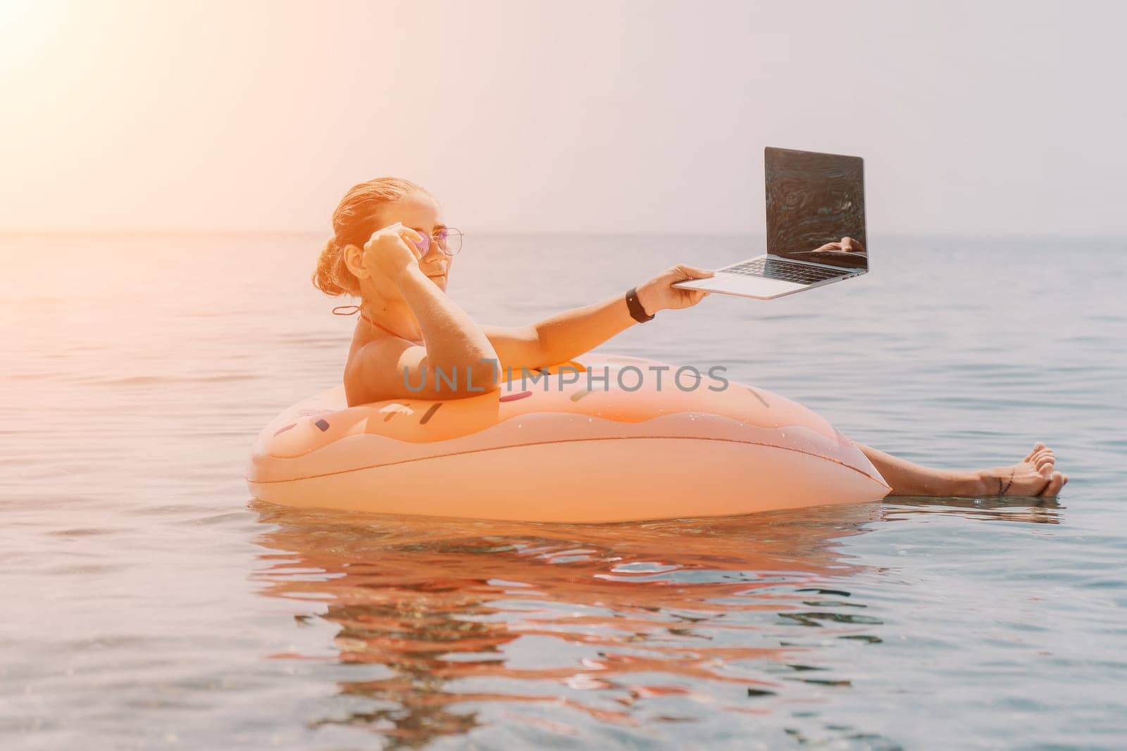 Woman freelancer works on laptop swimming in sea on pink inflatable ring. Pretty lady typing on computer while floating in the sea on inflatable donut at sunset. Freelance, remote work on vacation