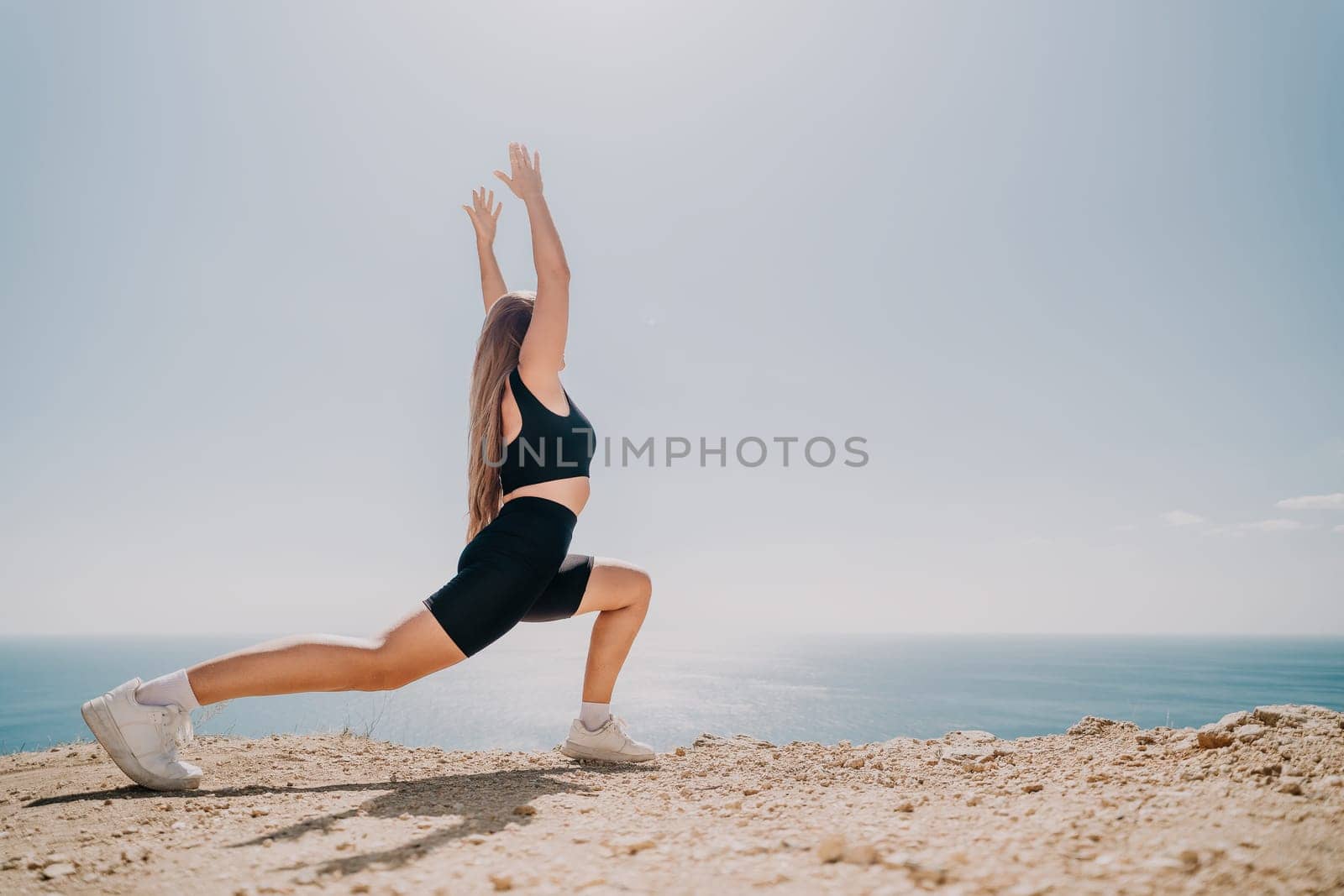 A woman wearing black workout clothes stretches her arms above her head in a yoga pose with the ocean in the background