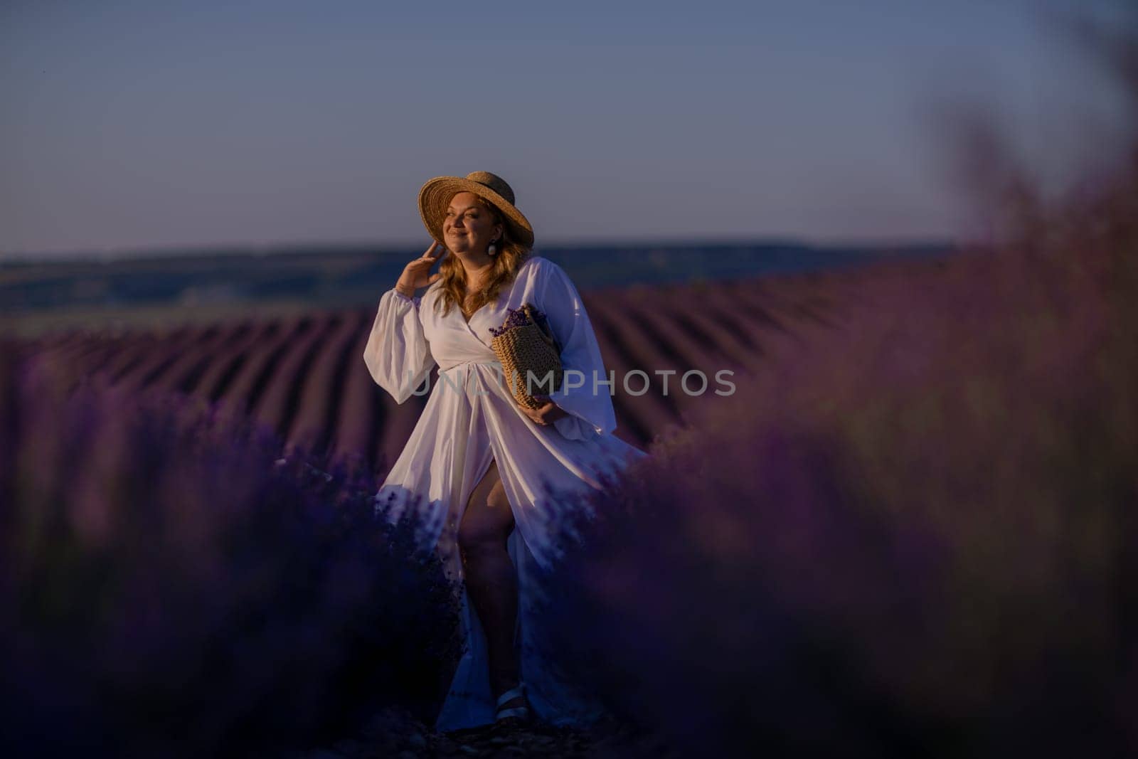 Woman poses in lavender field at sunset. Happy woman in white dress holds lavender bouquet. Aromatherapy concept, lavender oil, photo session in lavender.