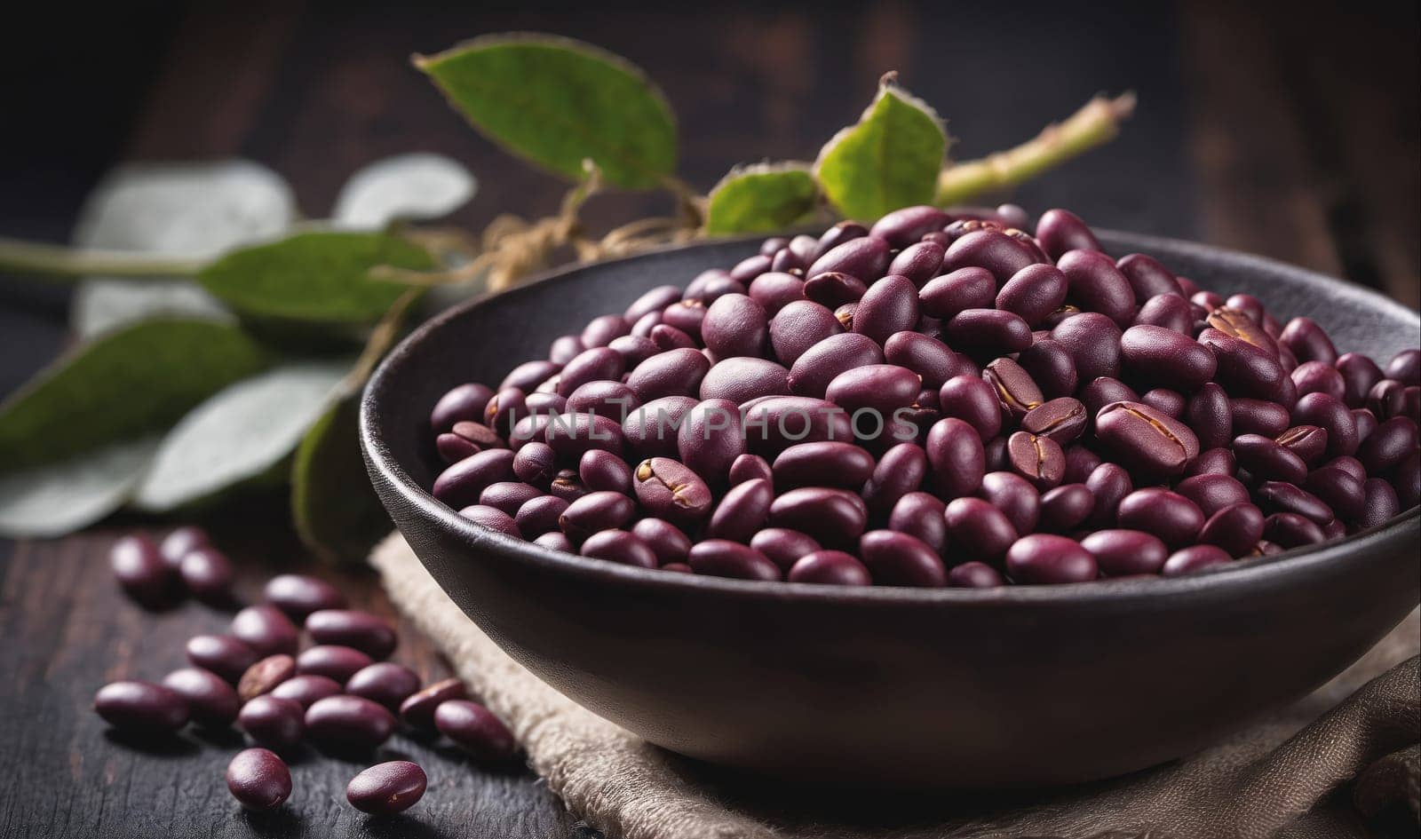 A bowl of red beans sits on a dark wooden surface. The beans are piled high in the bowl, which is black with a glossy finish. A few red beans are scattered on the surface of the table. Green leaves are visible behind the bowl, adding a touch of color to the scene.