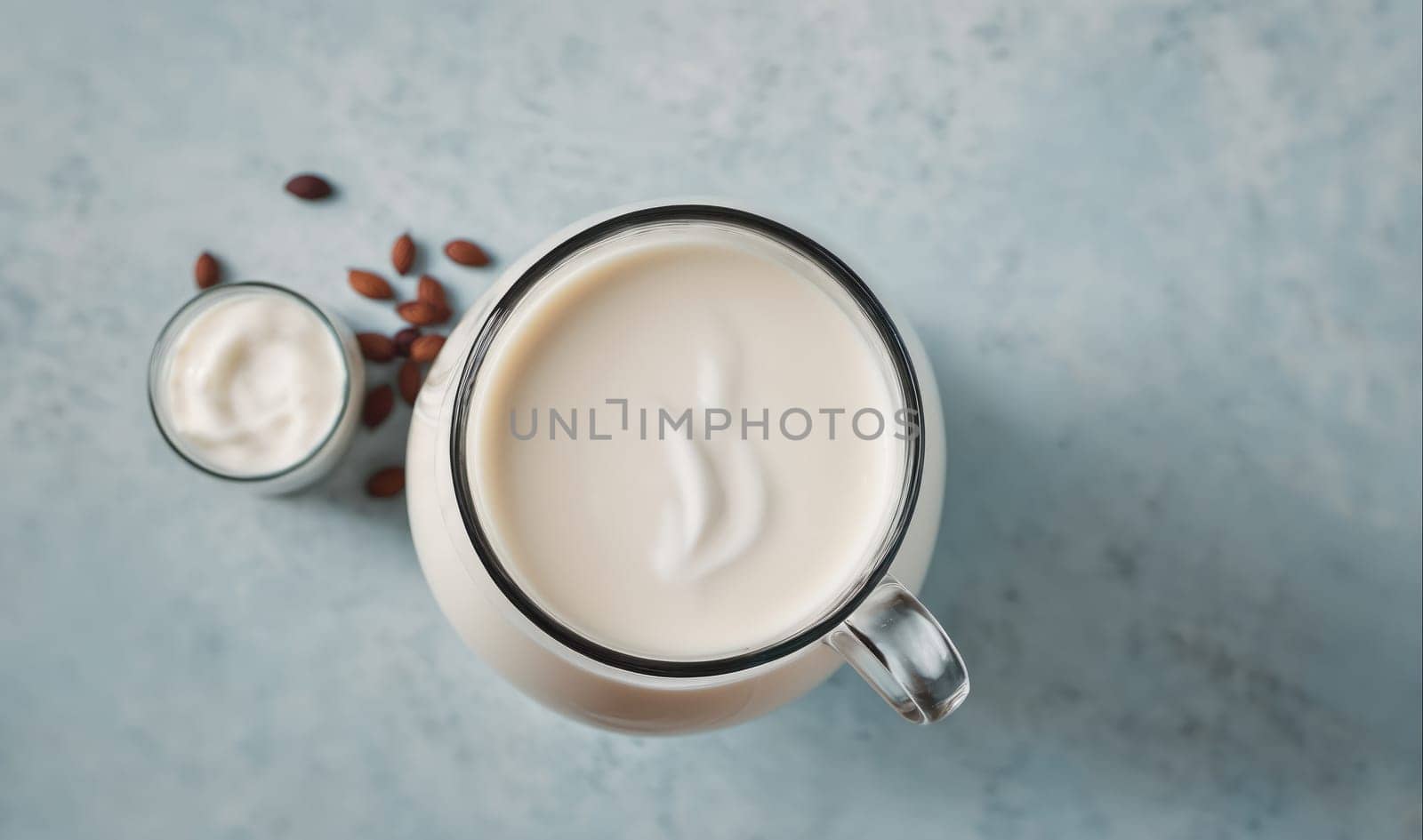 A pitcher and glass of almond milk are shown from a high angle on a blue countertop. Scattered almonds can be seen around the pitcher and glass.