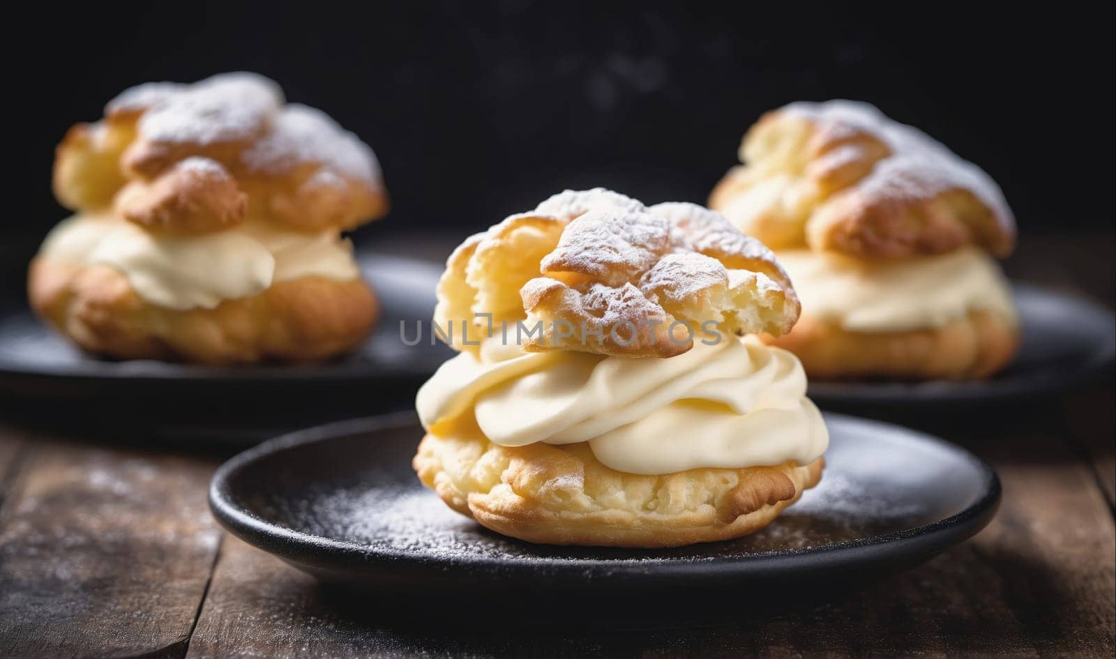 Three cream puffs, filled with whipped cream, are arranged on black plates. The pastries are dusted with powdered sugar and sit on a wooden surface.
