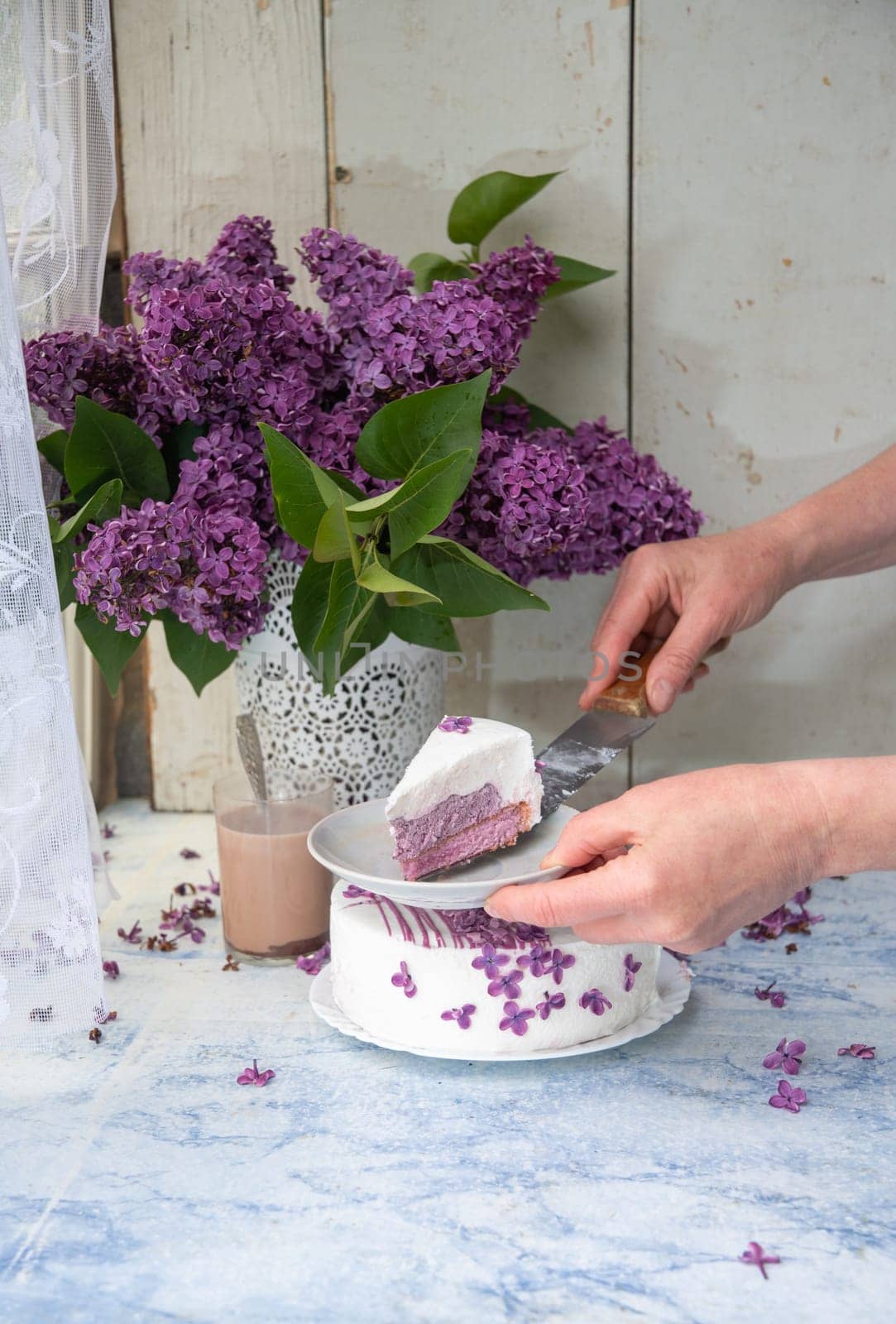 female hand cutting cake decorated with lilac flowers near vase with lilac branches on wooden table emphasizing floral and rustic lifestyle, delicate still life,