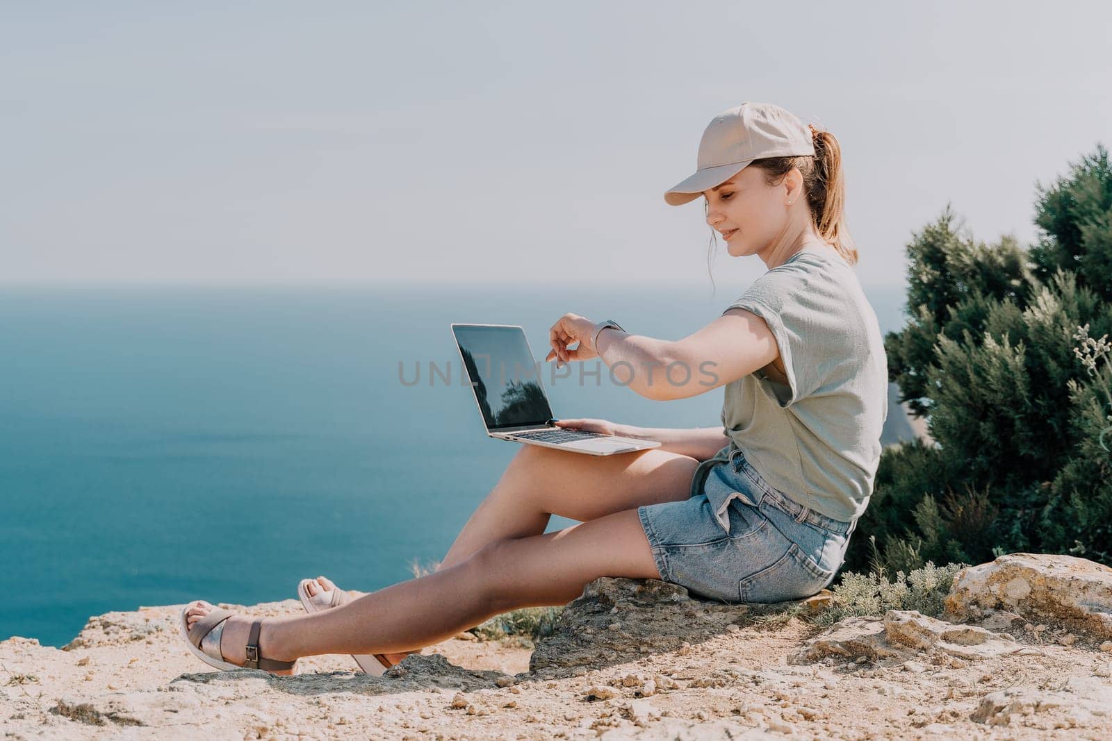 A young woman in a hat and casual clothes sits on a cliffside with a laptop, overlooking the ocean.