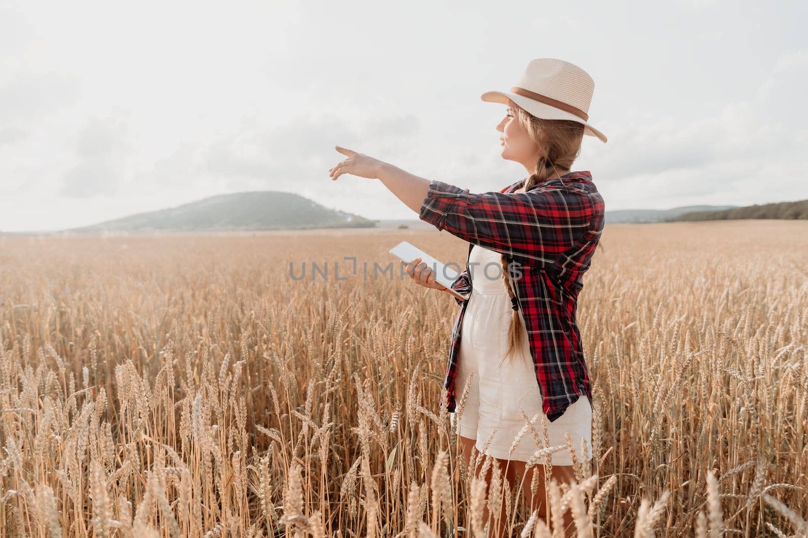 A young woman wearing a white hat and plaid shirt stands in a wheat field, pointing towards the horizon, holding a tablet.