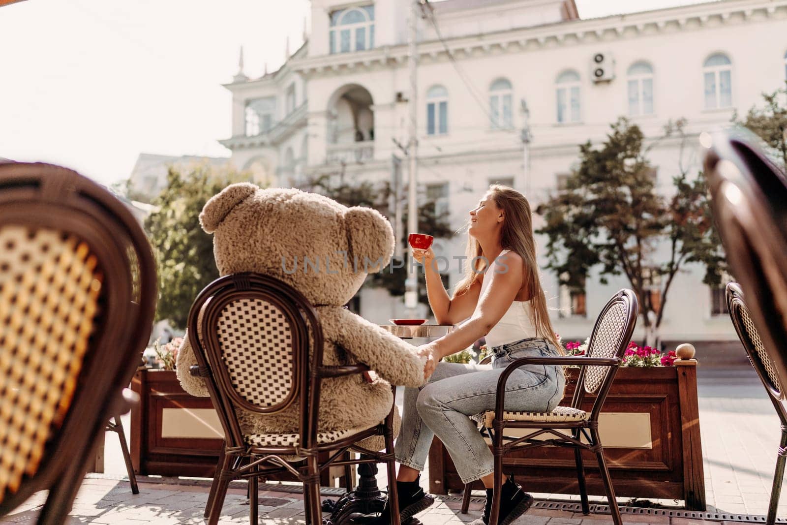 A woman sits cafe with a teddy bear next to her. The scene is set in a city with several chairs and tables around her. The woman is enjoying her time at the outdoor cafe