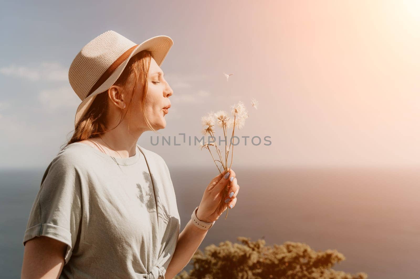 A young woman with red hair, wearing a hat, blows on a bunch of dandelions, while standing against a bright, sunny sky.