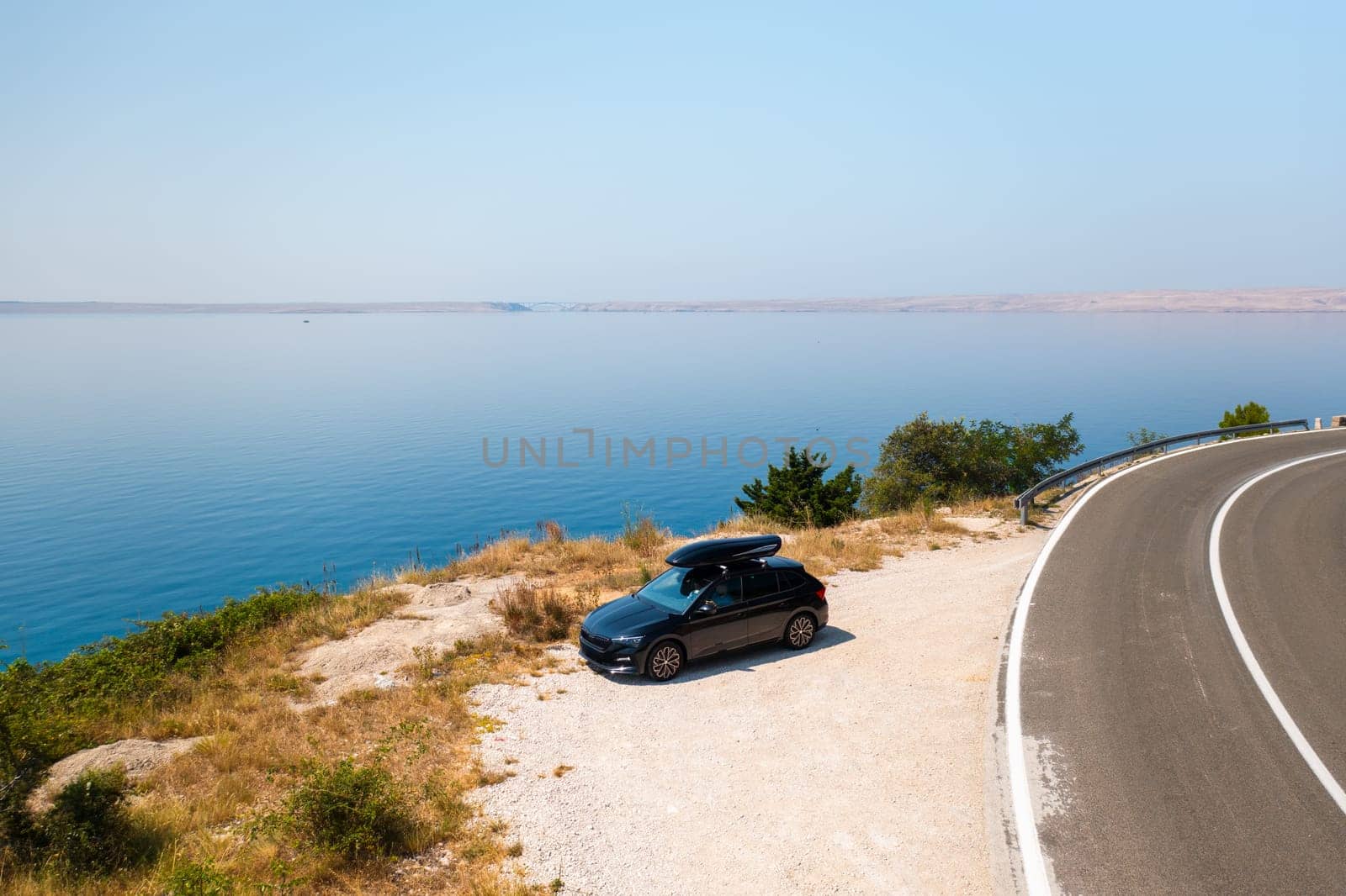 Car with a roof box, parked beside a narrow, winding road on a green cliff, with the azure sea