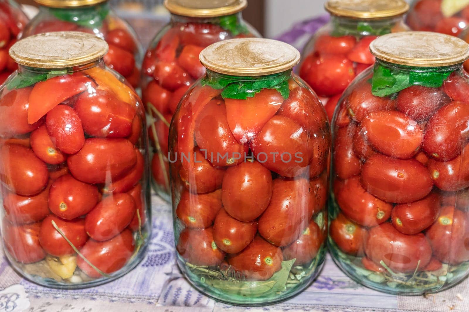 Vibrant jars filled with red tomatoes are neatly arranged on a table, showcasing the process of home canning for preservation. Fresh herbs accentuate the jars, highlighting freshness.