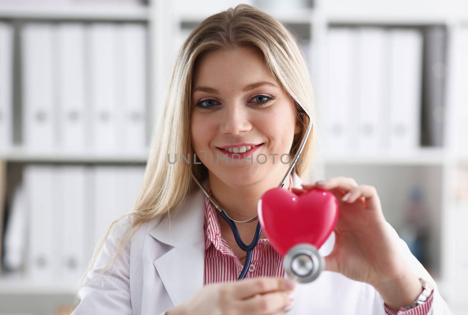 Beautiful smiling blond female doctor hold in arms red toy heart closeup. Cardio therapeutist student education CPR 911 life save physician make cardiac physical pulse rate measure arrhythmia