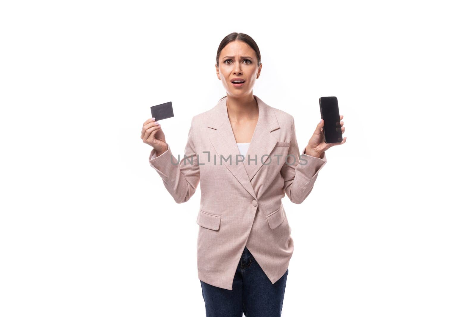 pleasant young woman with black hair in a jacket holding a credit card and a smartphone on a white background with copy space by TRMK