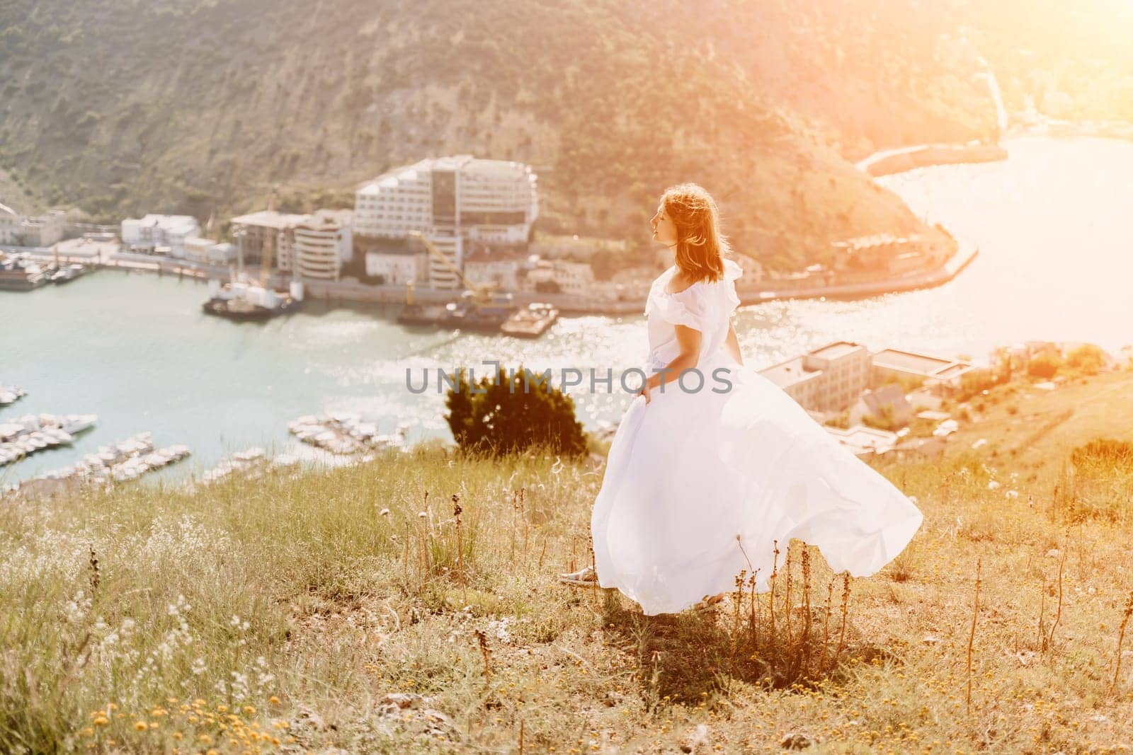 Happy woman in a white dress and hat stands on a rocky cliff above the sea, with the beautiful silhouette of hills in thick fog in the background. by Matiunina