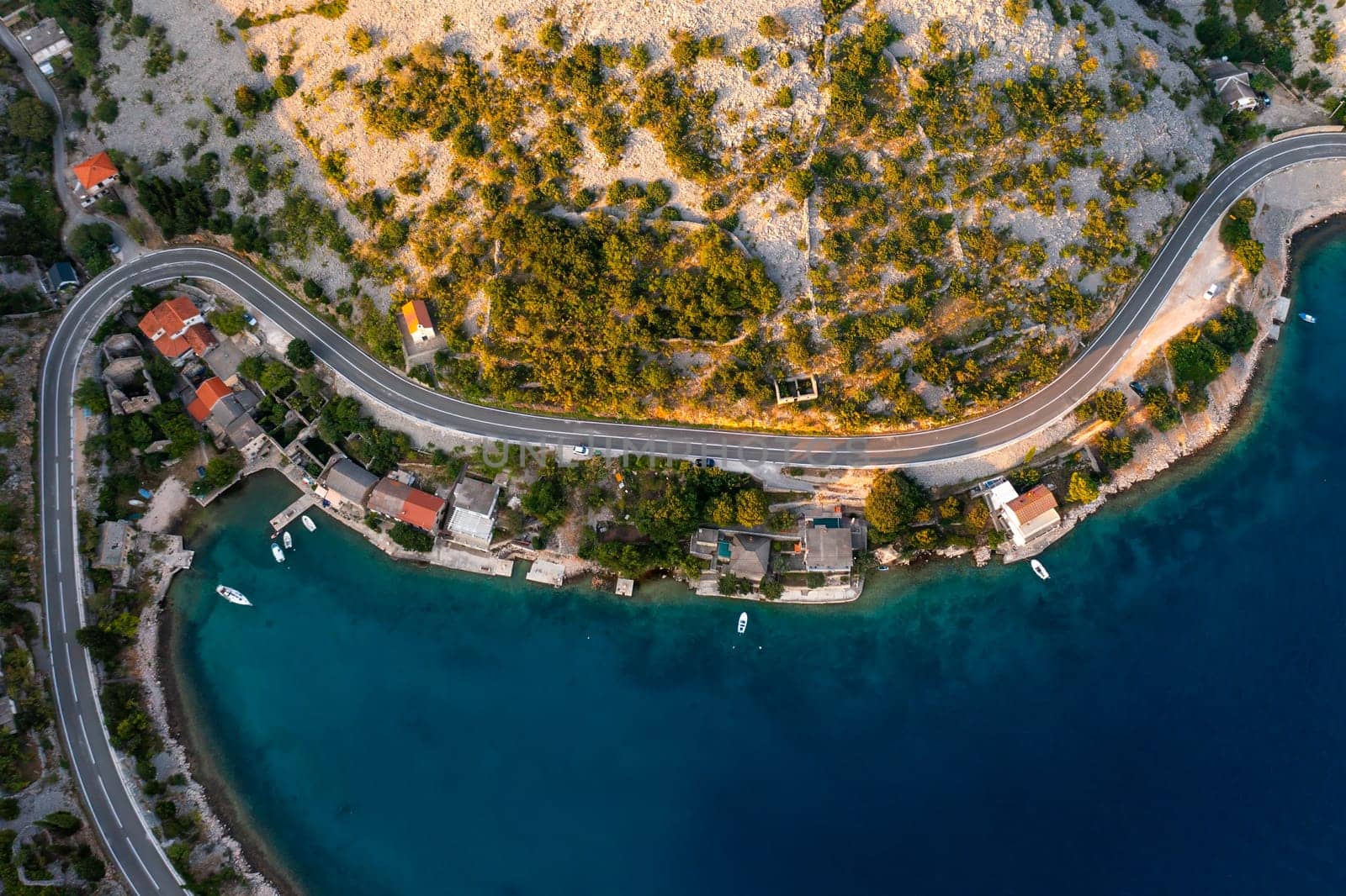 Aerial view of a coastal village with winding road and clear blue water in the afternoon sun.