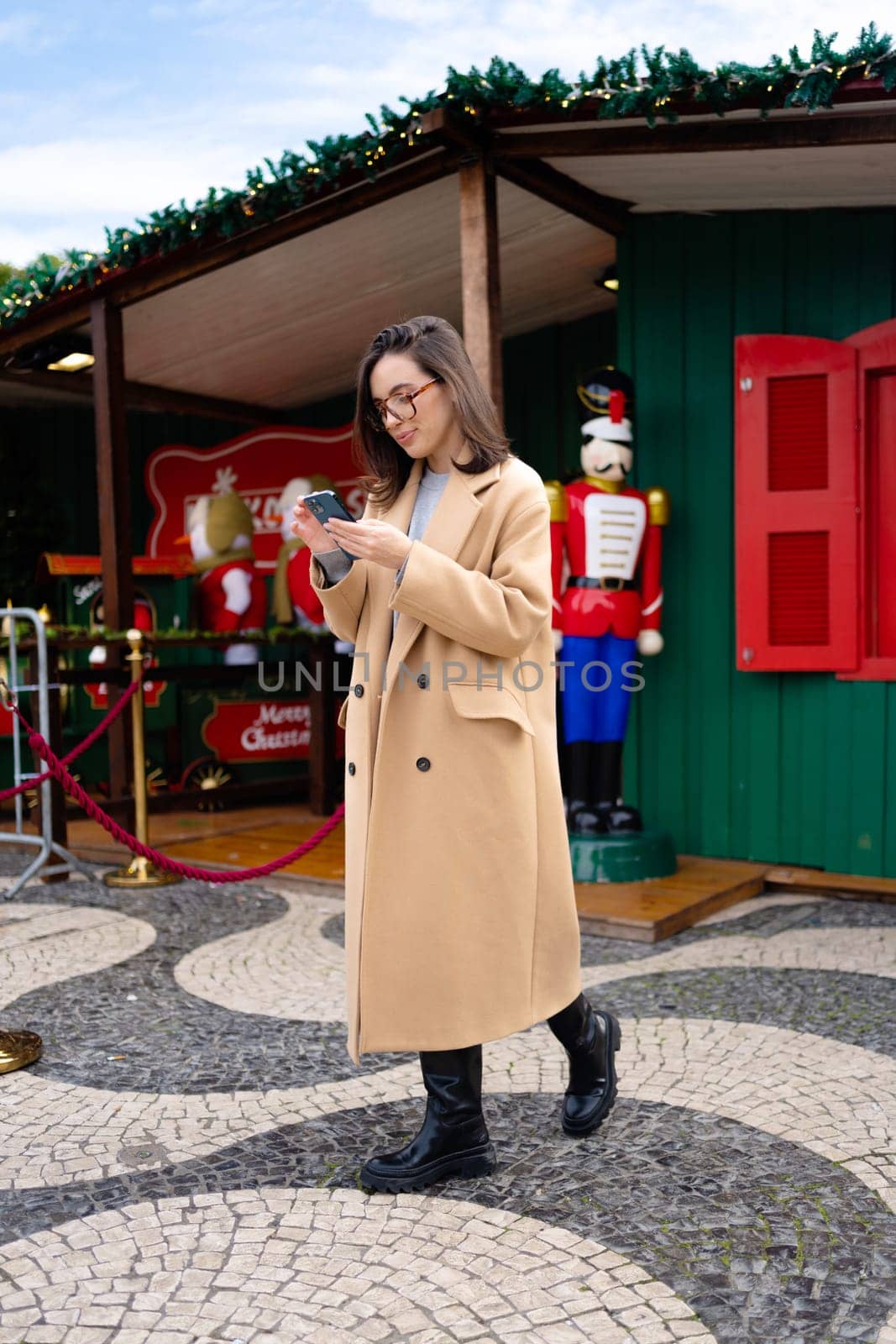 Woman in glasses using mobile phone at day in the city. Happy woman using smartphone dressed stylish trench coat looking device screen. Vertical photo full length