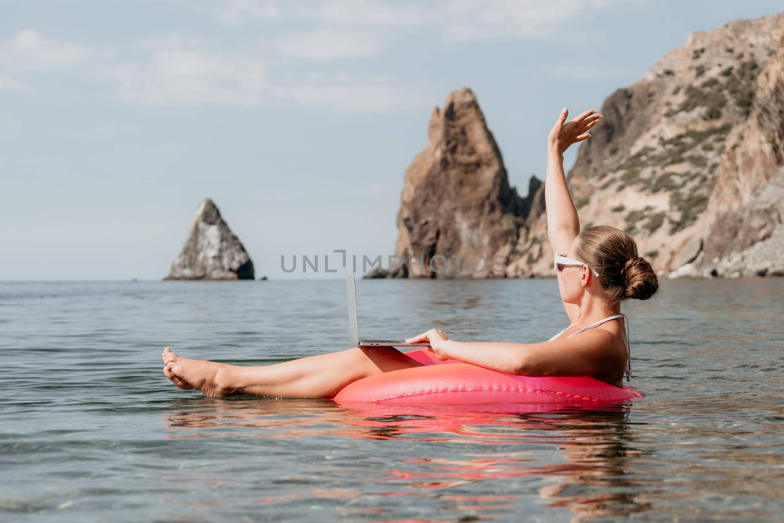Woman freelancer works on laptop swimming in sea on pink inflatable ring. Pretty lady typing on computer while floating in the sea on inflatable donut at sunset. Freelance, remote work on vacation
