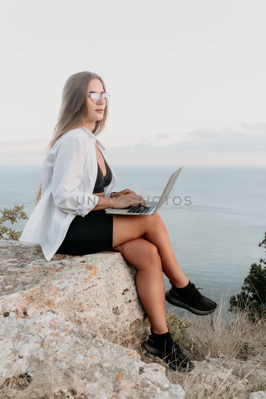 Digital nomad, woman in the hat, a business woman with a laptop sits on the rocks by the sea during sunset, makes a business transaction online from a distance. Freelance, remote work on vacation.