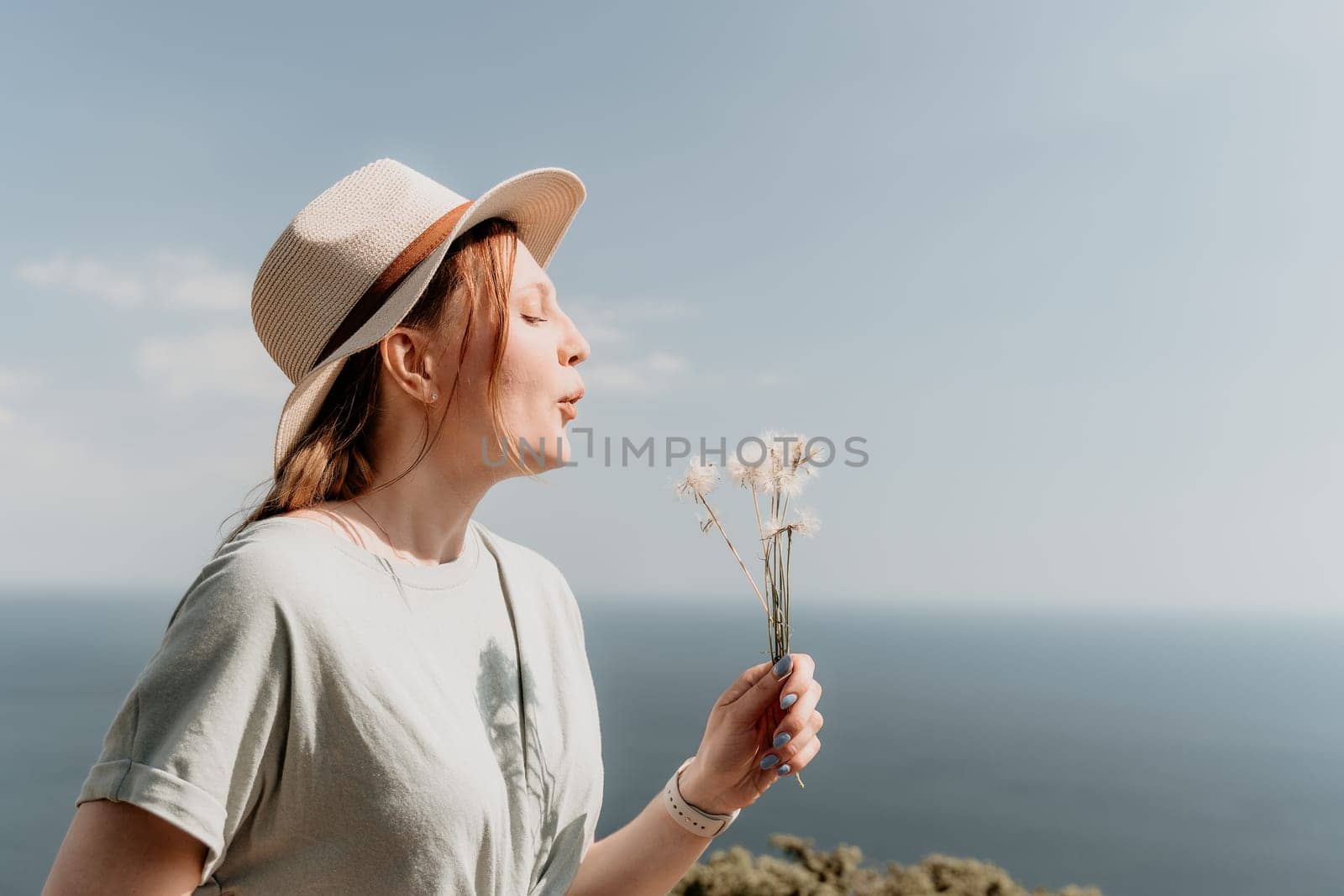 A young woman wearing a hat blows on a dandelion while standing on a cliff overlooking the ocean.