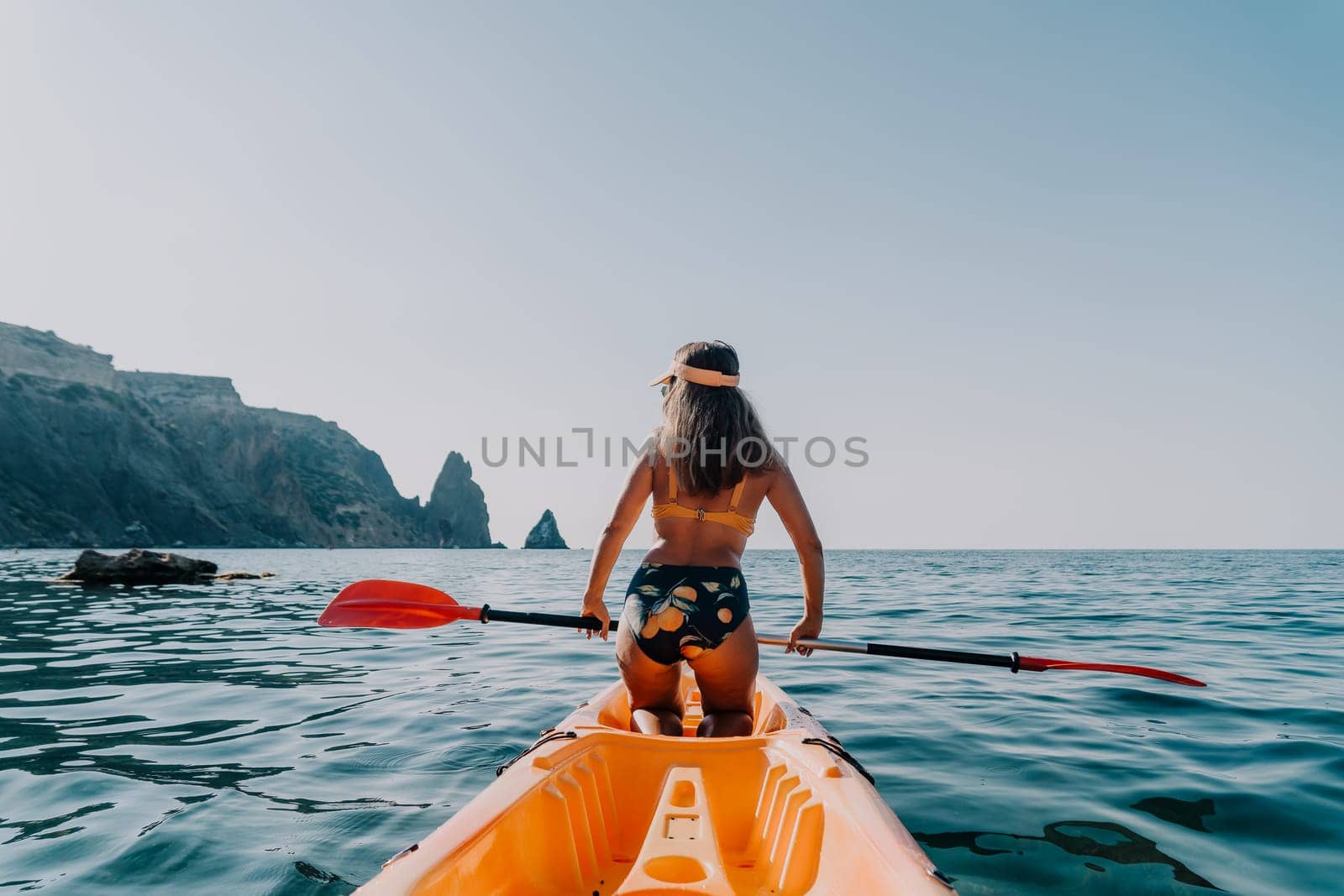 A woman in a bikini paddles a kayak towards rocky cliffs in the distance. The sea is calm and the sky is blue.