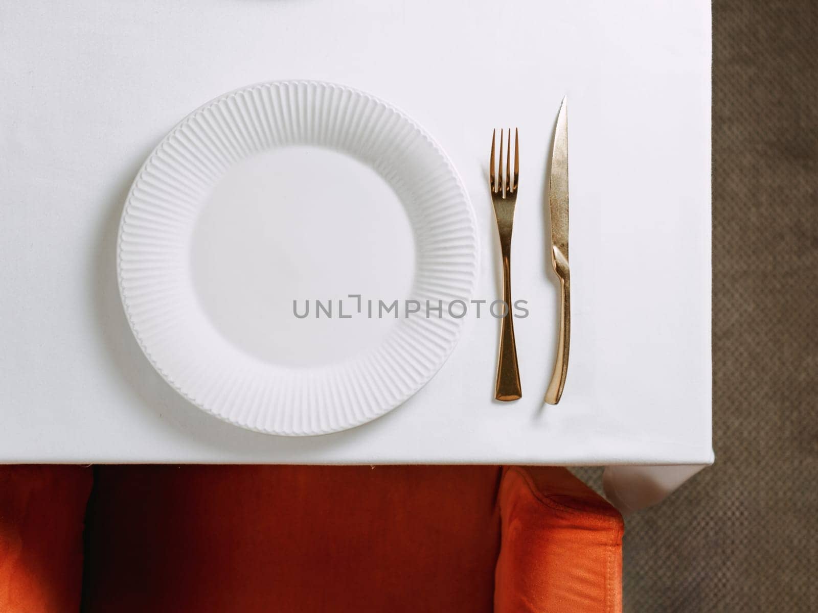 Top view of table in restaurant or dinning room with white round empty plate and gold cutlery. White tablecloth, red chair and table setting as background for your text or design. Copy space