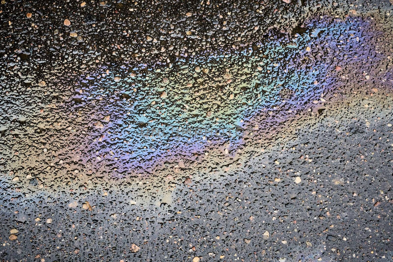 A top-down view of a reflective puddle of oil or gasoline on saturated asphalt.