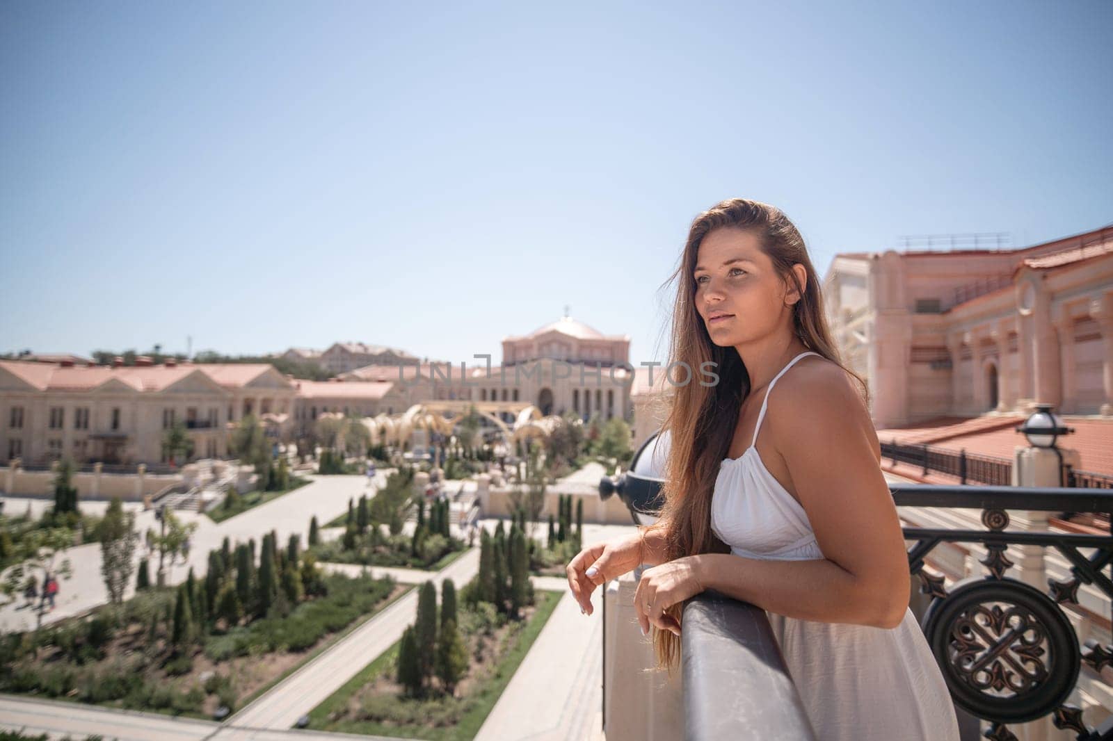 A woman in a white dress stands on a balcony overlooking a sprawling cityscape. Buildings with terracotta roofs and manicured gardens are visible in the distance. The sky is clear and blue.