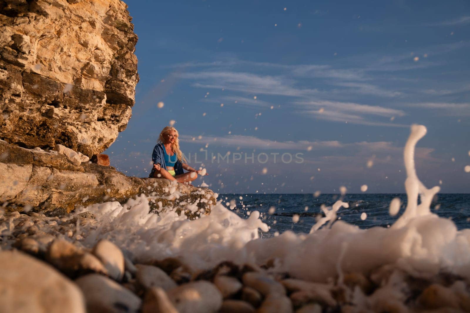 A woman meditates on a rocky shore, facing a crashing wave. The setting sun casts warm light on the scene.